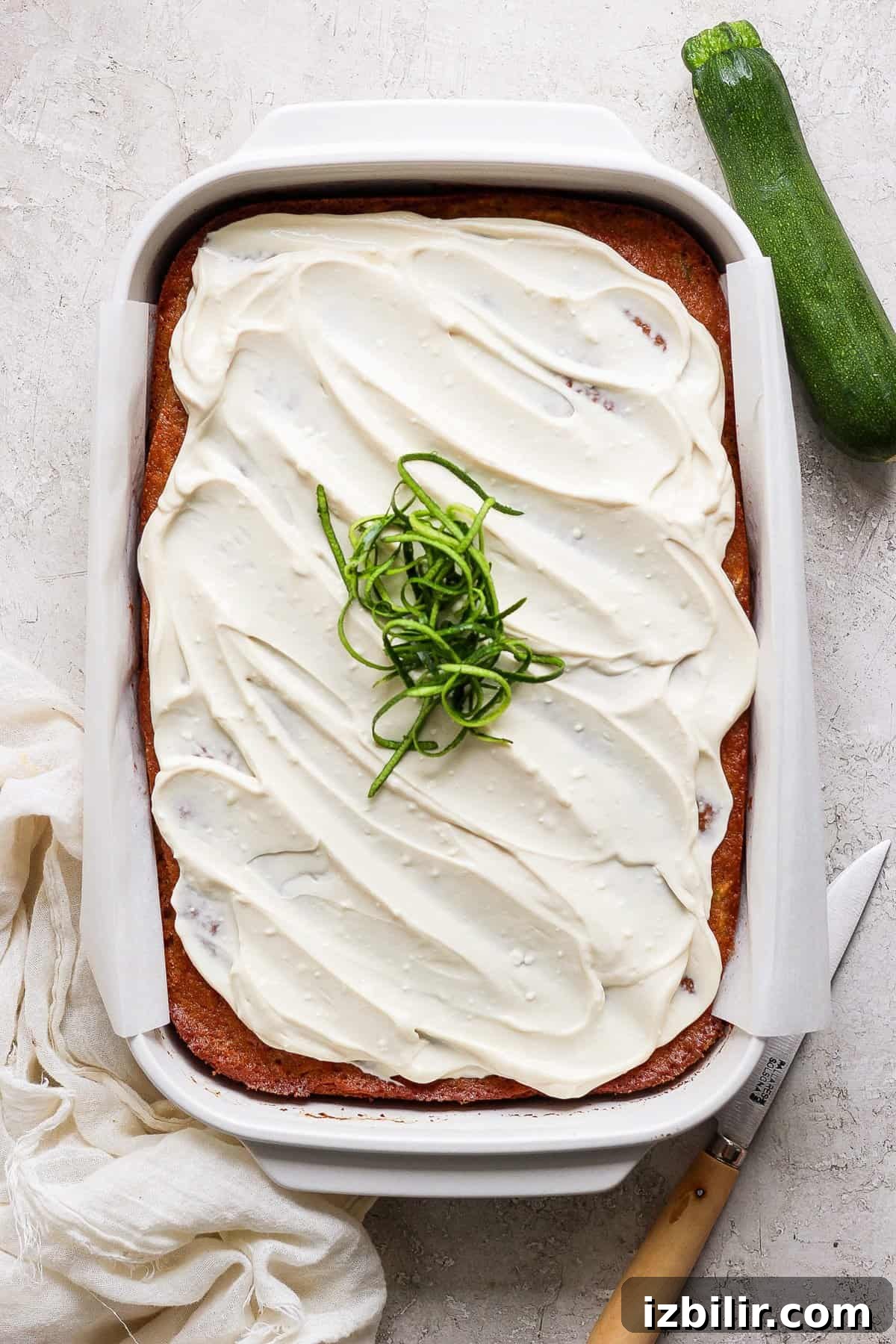 Moist Low-Sugar Zucchini Cake Recipe 4 A frosted sheet cake garnished with spiralized zucchini sits in a rectangular baking dish. A knife, cloth, and a whole zucchini are placed beside the dish.