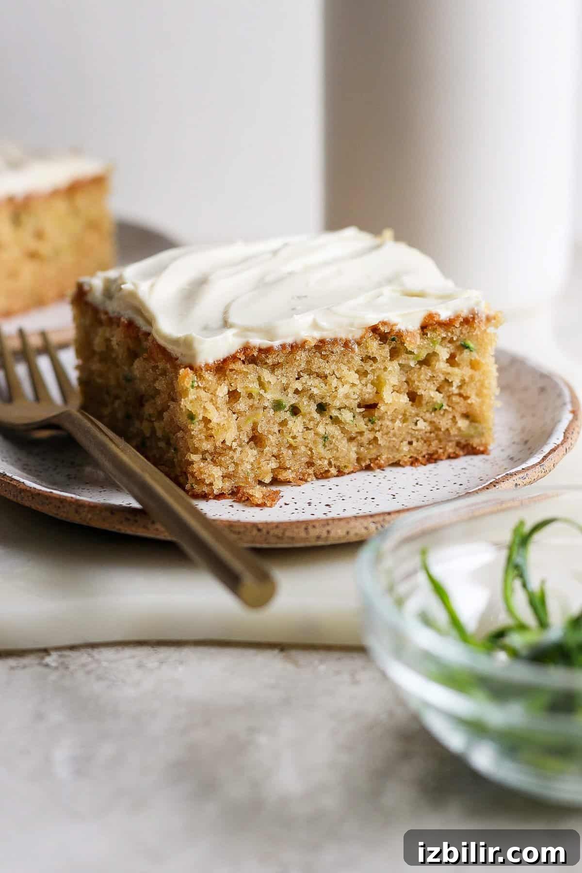 Moist Low-Sugar Zucchini Cake Recipe 5 A slice of frosted zucchini cake on a plate with a fork beside it. A bowl with green garnish is in the foreground.