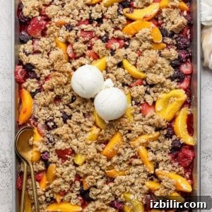 A rectangular baking tray filled with a mixed berry and peach crumble topped with two scoops of vanilla ice cream. A gold spoon rests on the left side of the tray.