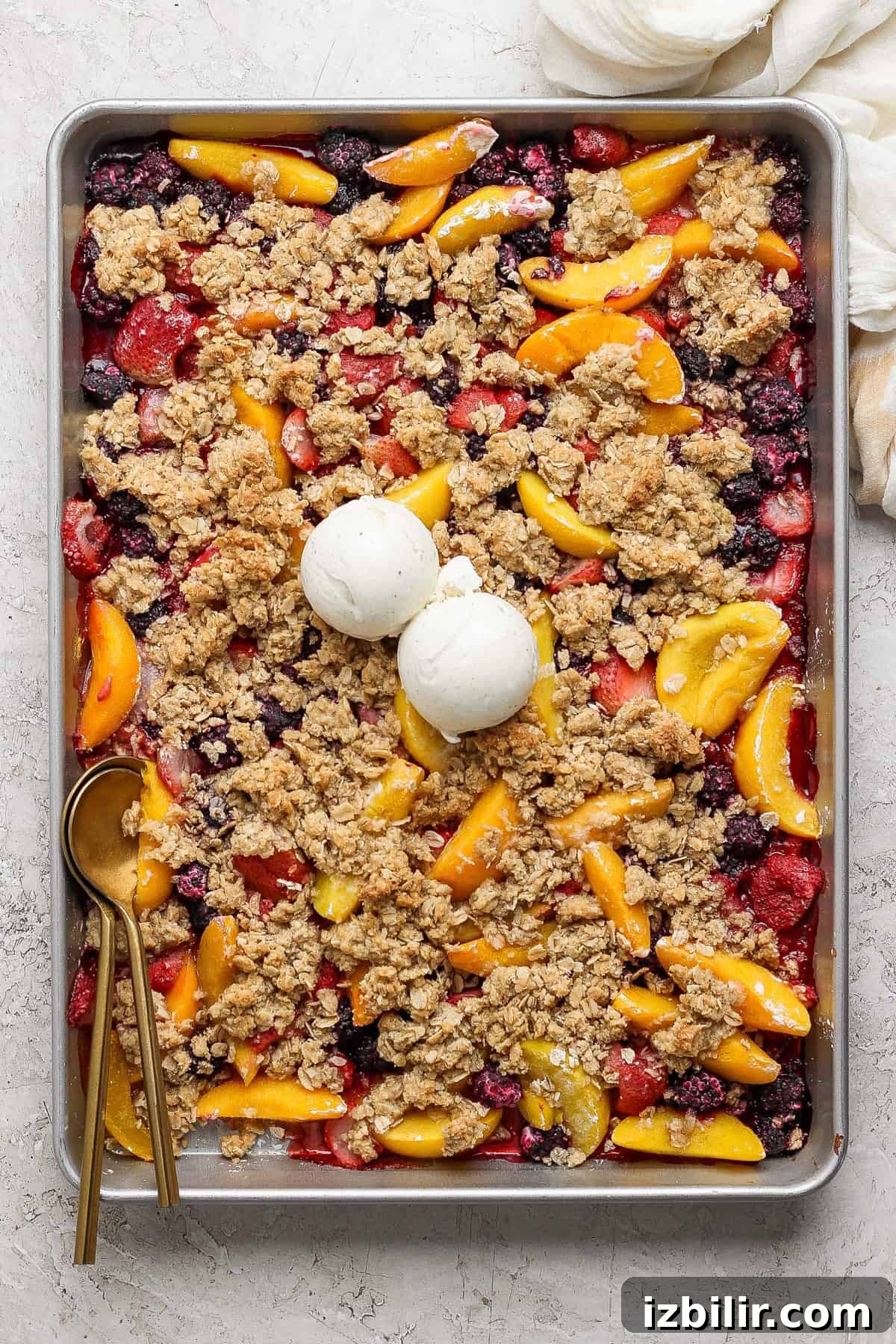 A rectangular baking tray filled with a mixed berry and peach crumble topped with two scoops of vanilla ice cream. A gold spoon rests on the left side of the tray.