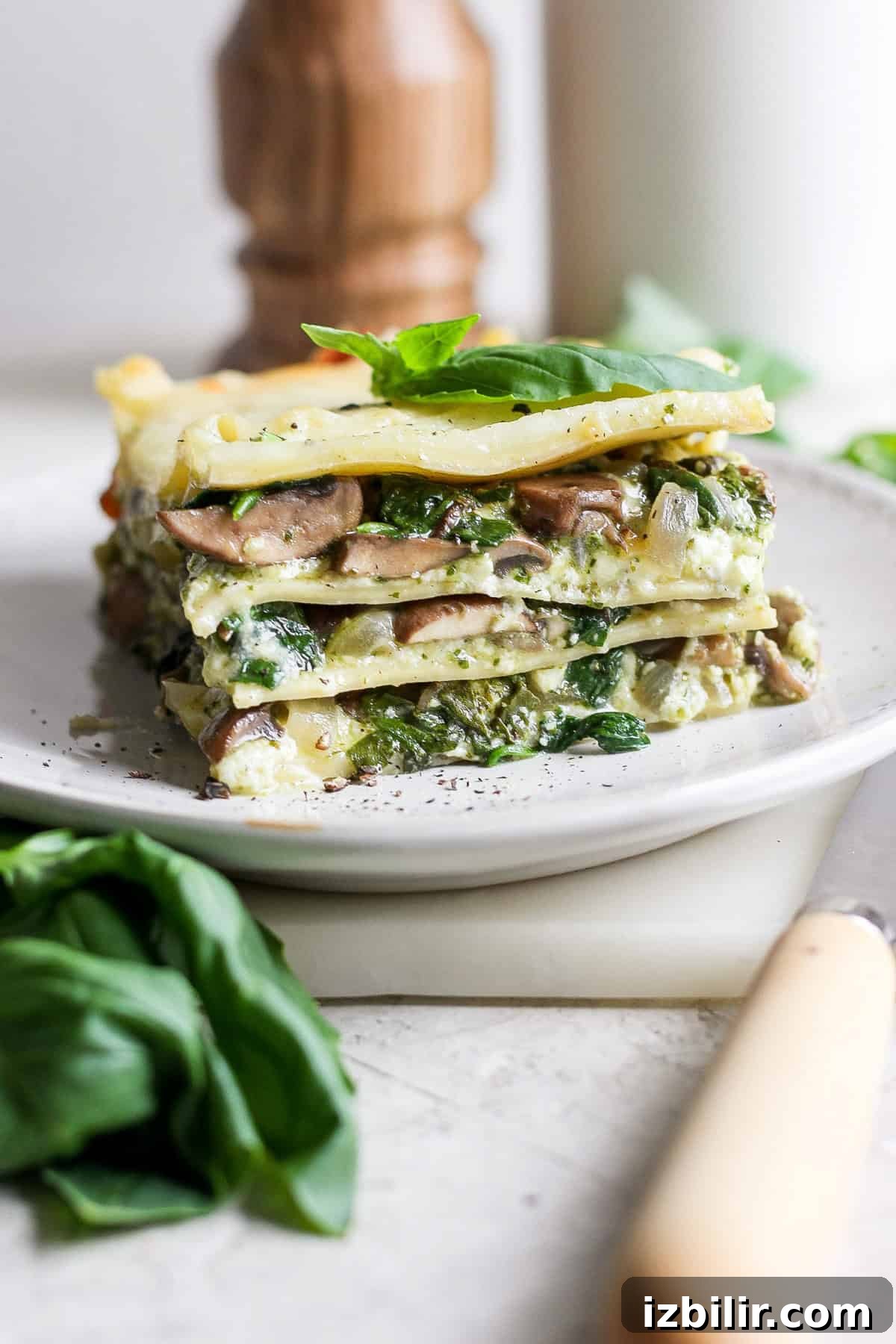 Pesto Lasagna Recipe with Fresh Basil and Mozzarella 2 A slice of vegetarian lasagna with visible layers of pasta, spinach, mushrooms, and cheese on a plate, garnished with fresh basil leaves.