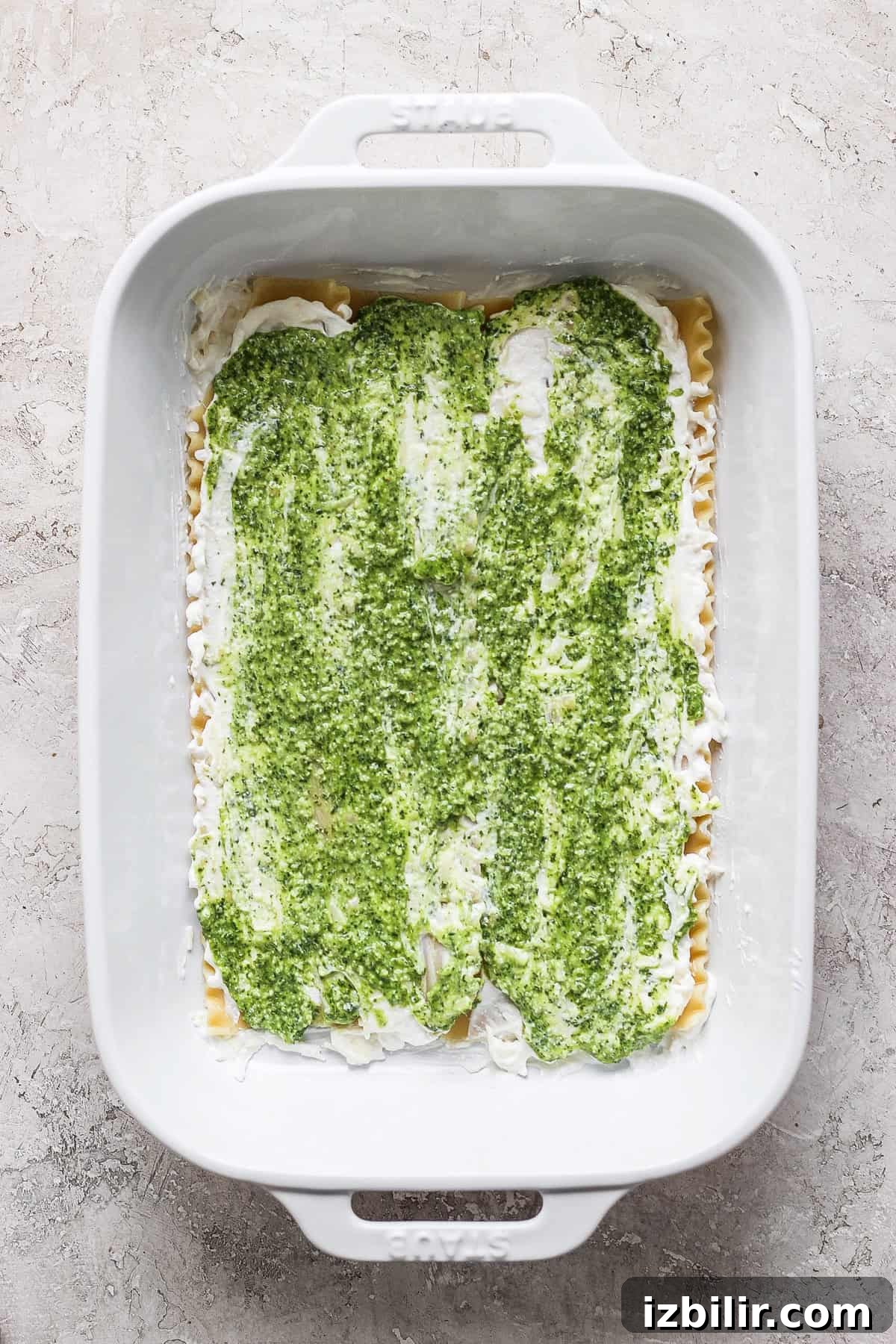 Pesto Lasagna Recipe with Fresh Basil and Mozzarella 4 A white baking dish containing an uncooked layered green and white lasagna against a textured light gray surface.