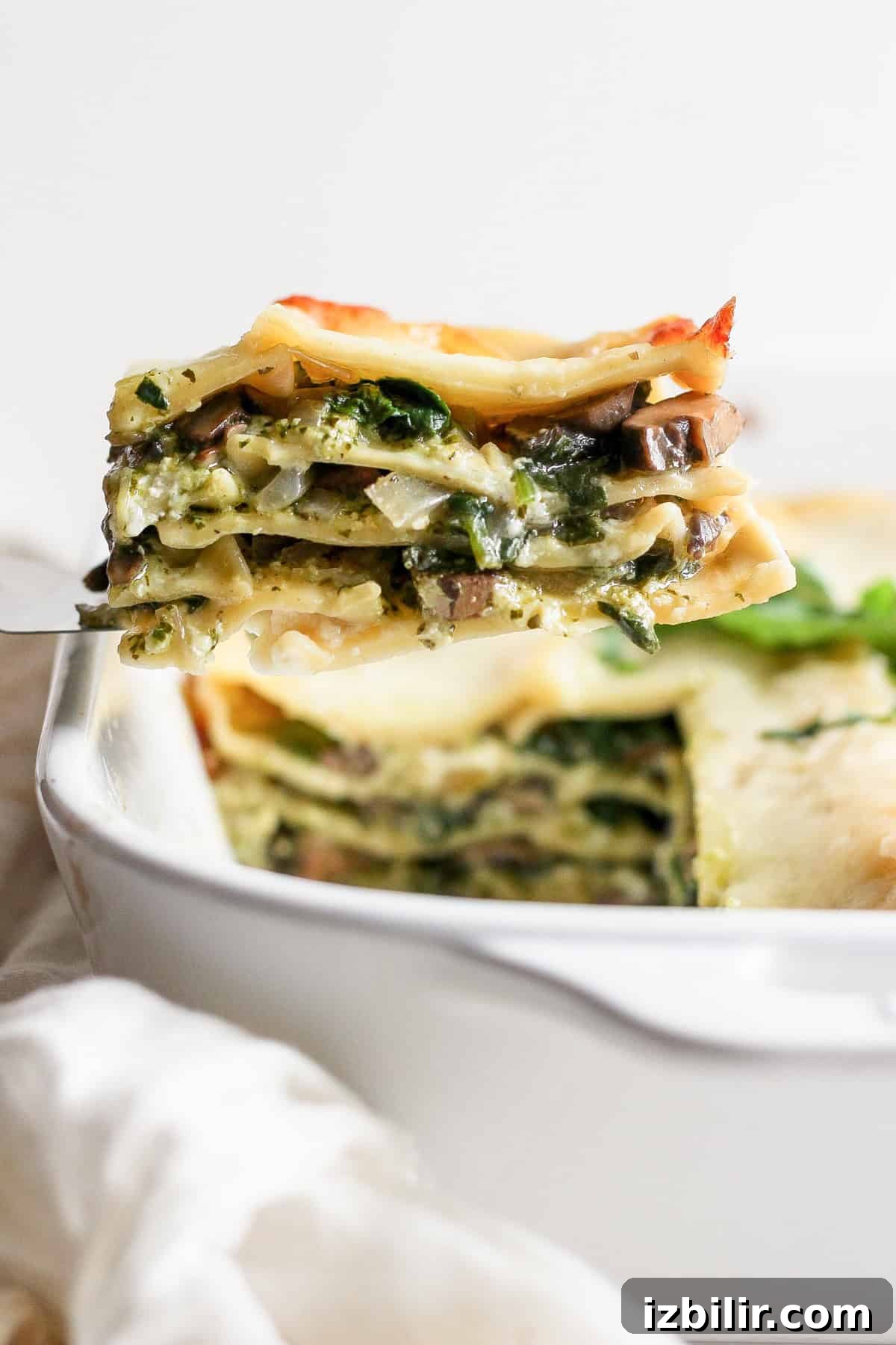 Pesto Lasagna Recipe with Fresh Basil and Mozzarella 7 A close-up of a slice of vegetable lasagna being lifted from a white baking dish.