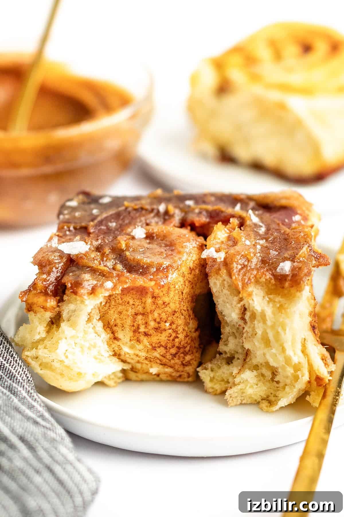 Caramel Glazed Apple Sticky Buns Recipe 2 Close-up of a cinnamon roll with glaze and a sprinkle of salt on a white plate. A partially eaten cinnamon roll and a dish with more glaze are in the background.