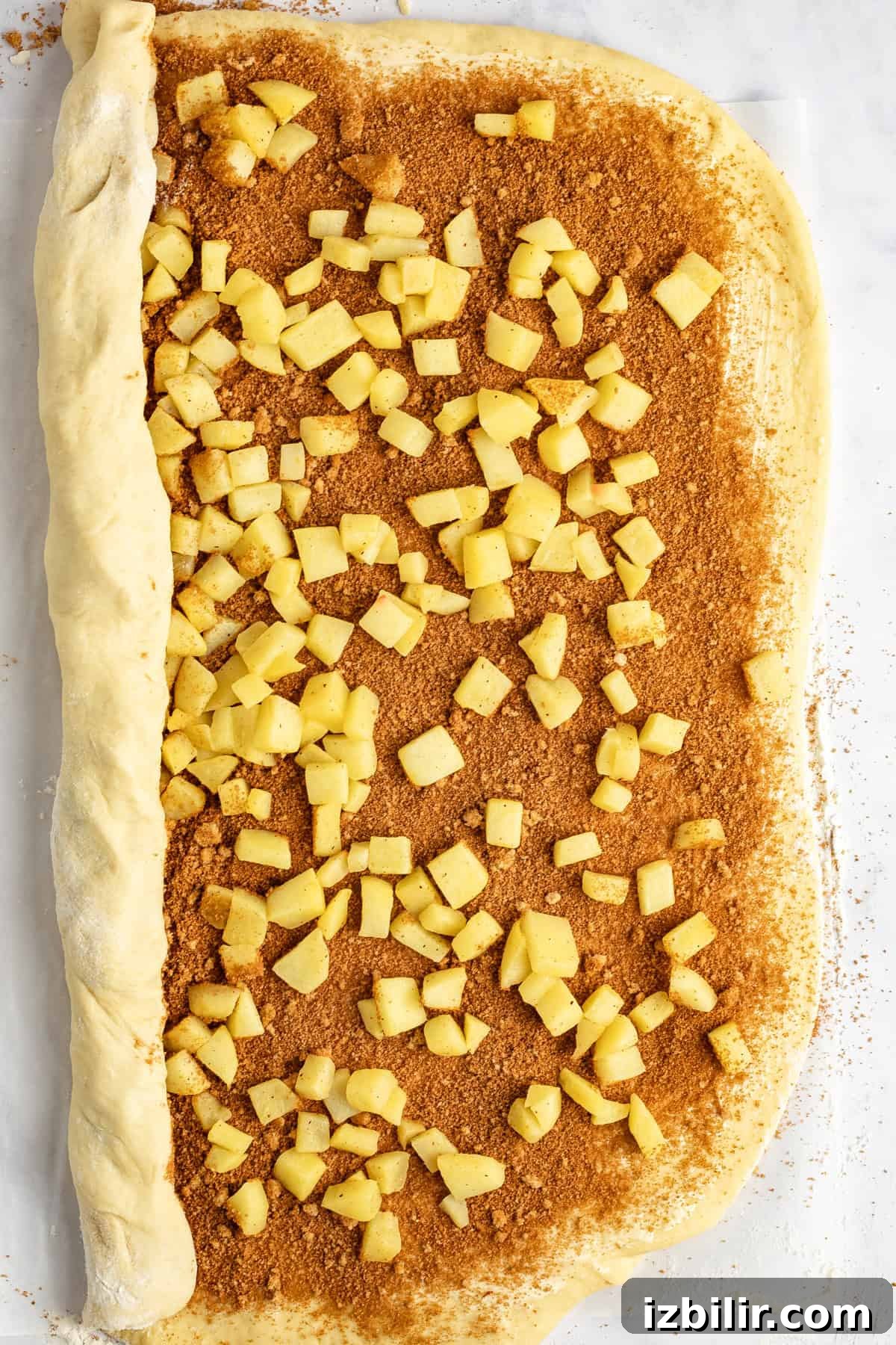 Caramel Glazed Apple Sticky Buns Recipe 4 Unbaked cinnamon roll dough topped with diced apples and cinnamon sugar, partially rolled up on a light surface.