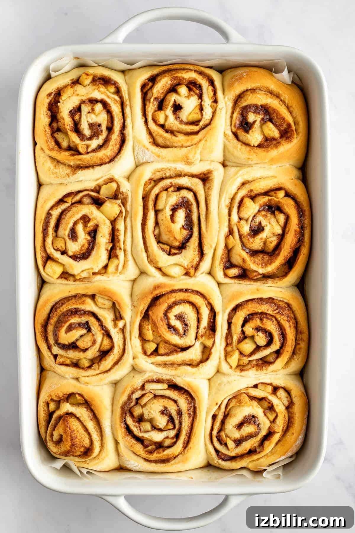 Caramel Glazed Apple Sticky Buns Recipe 5 A rectangular baking dish filled with twelve freshly baked cinnamon rolls, each with a visible swirl pattern, on a white surface.