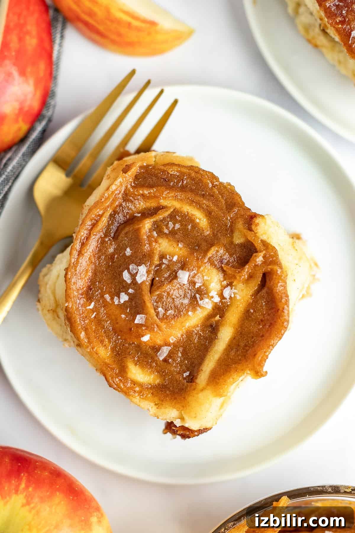 Caramel Glazed Apple Sticky Buns Recipe 6 A single cinnamon roll with frosting and a sprinkle of sea salt is on a white plate with a gold fork. Slices of a red apple are partially visible in the background.