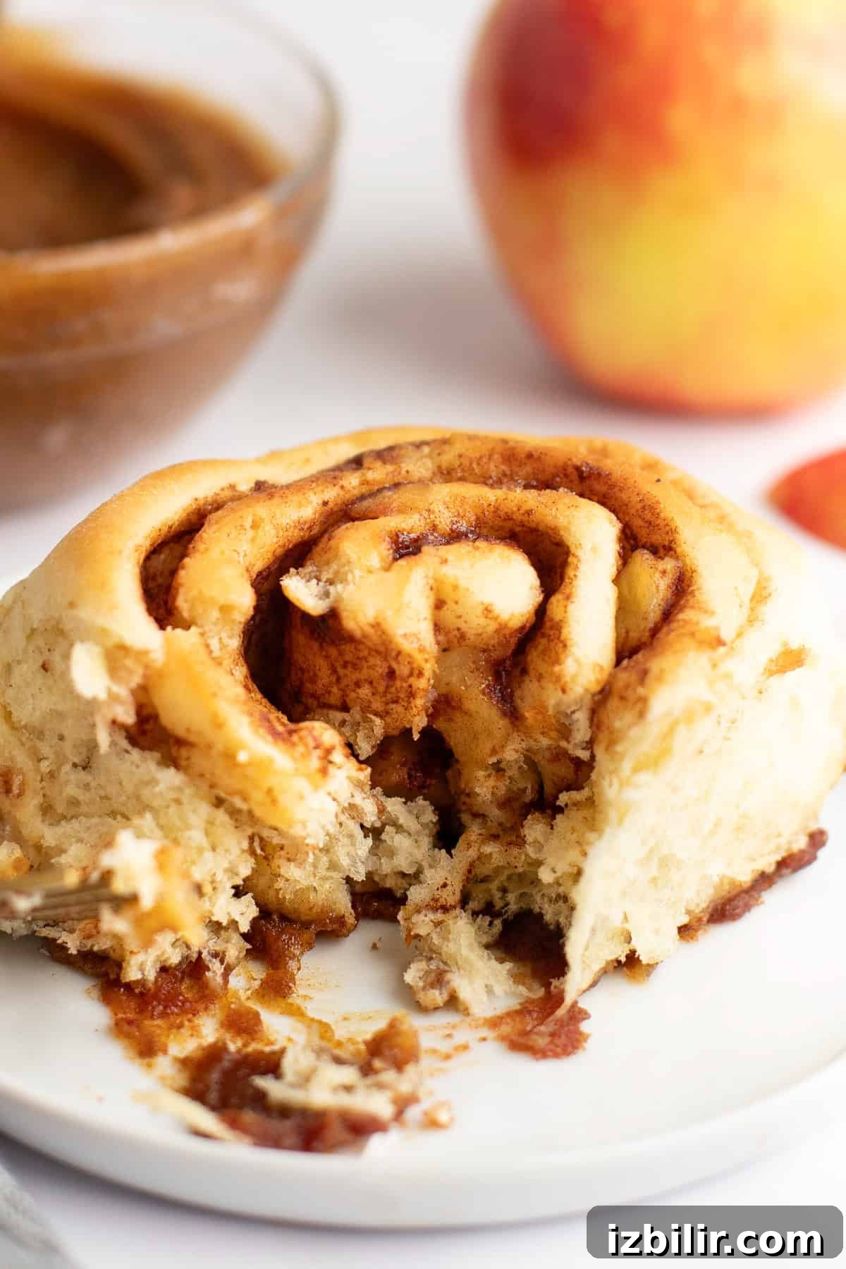 Caramel Glazed Apple Sticky Buns Recipe 7 A partially eaten cinnamon roll on a white plate with a bowl of filling and an apple in the background.