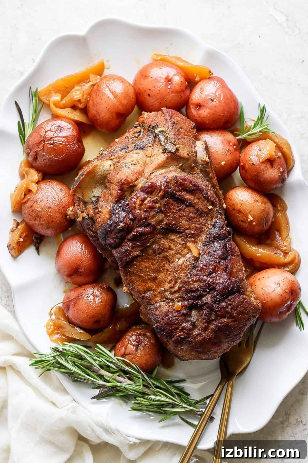 A roasted pork shoulder is served on a white plate, accompanied by red potatoes, onions, and garnished with rosemary sprigs.