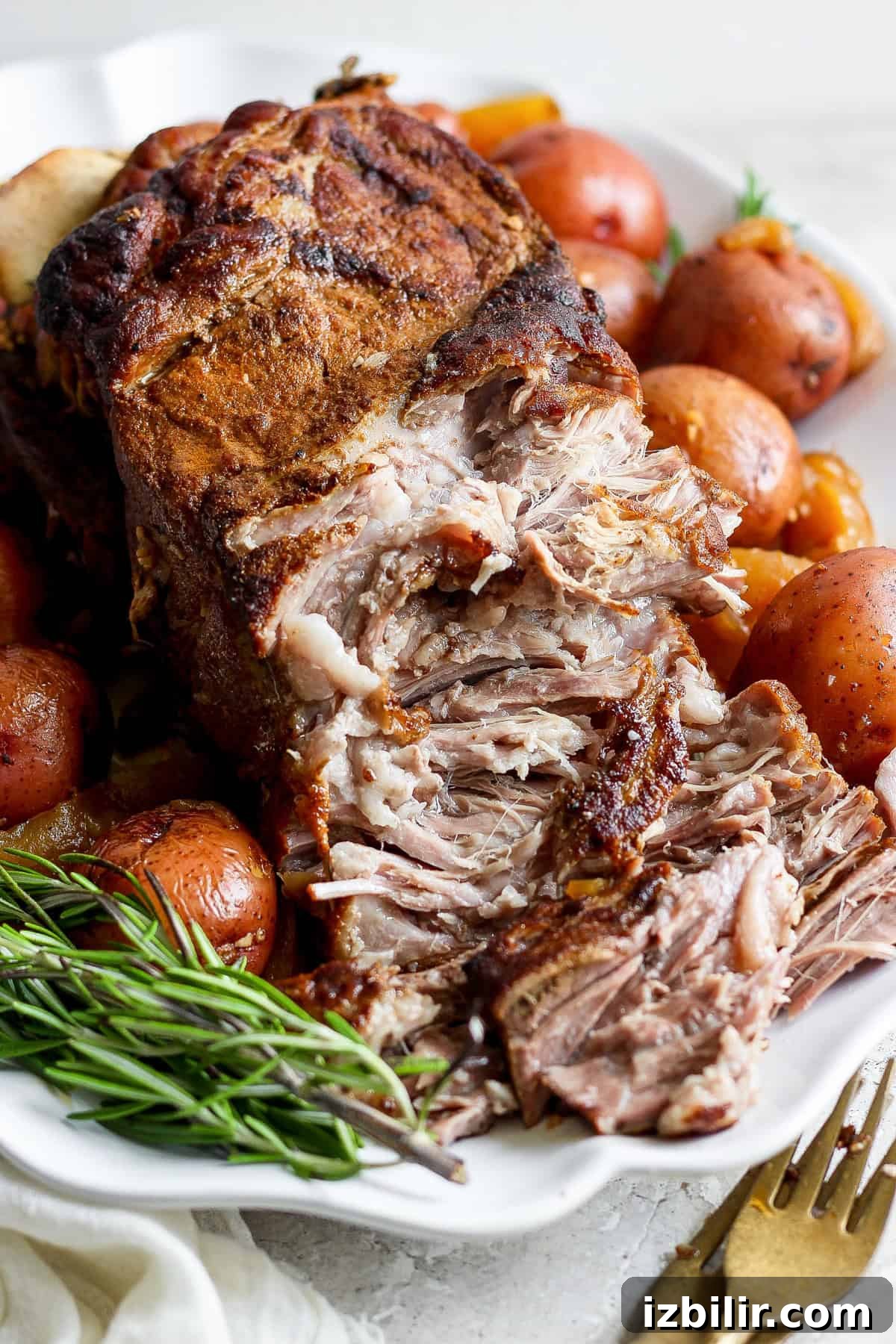 A close-up of a seasoned, cooked roast surrounded by whole red potatoes and fresh rosemary sprigs on a white platter.