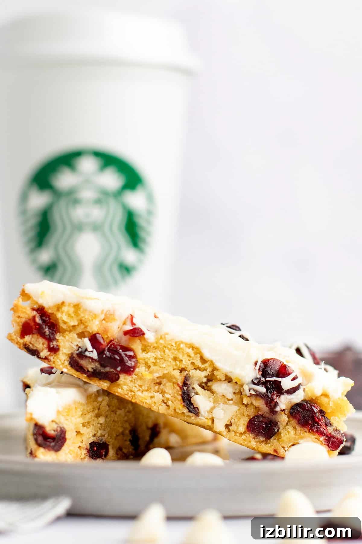 Two triangular slices of a white chocolate cranberry bar are stacked on a plate in front of a white Starbucks coffee cup.