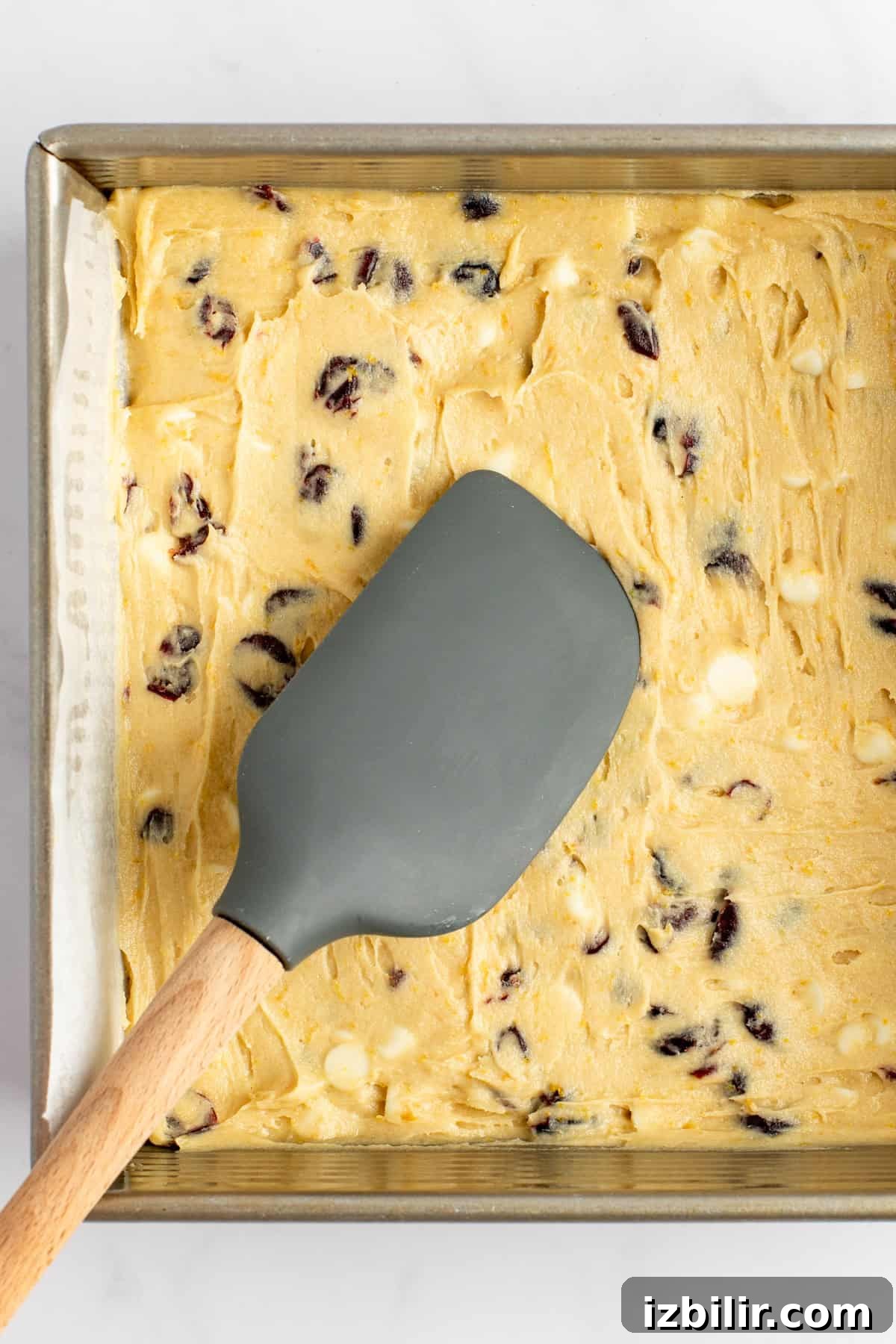 A baking tray filled with cookie dough containing cranberry and white chocolate chips, with a grey spatula resting on top.