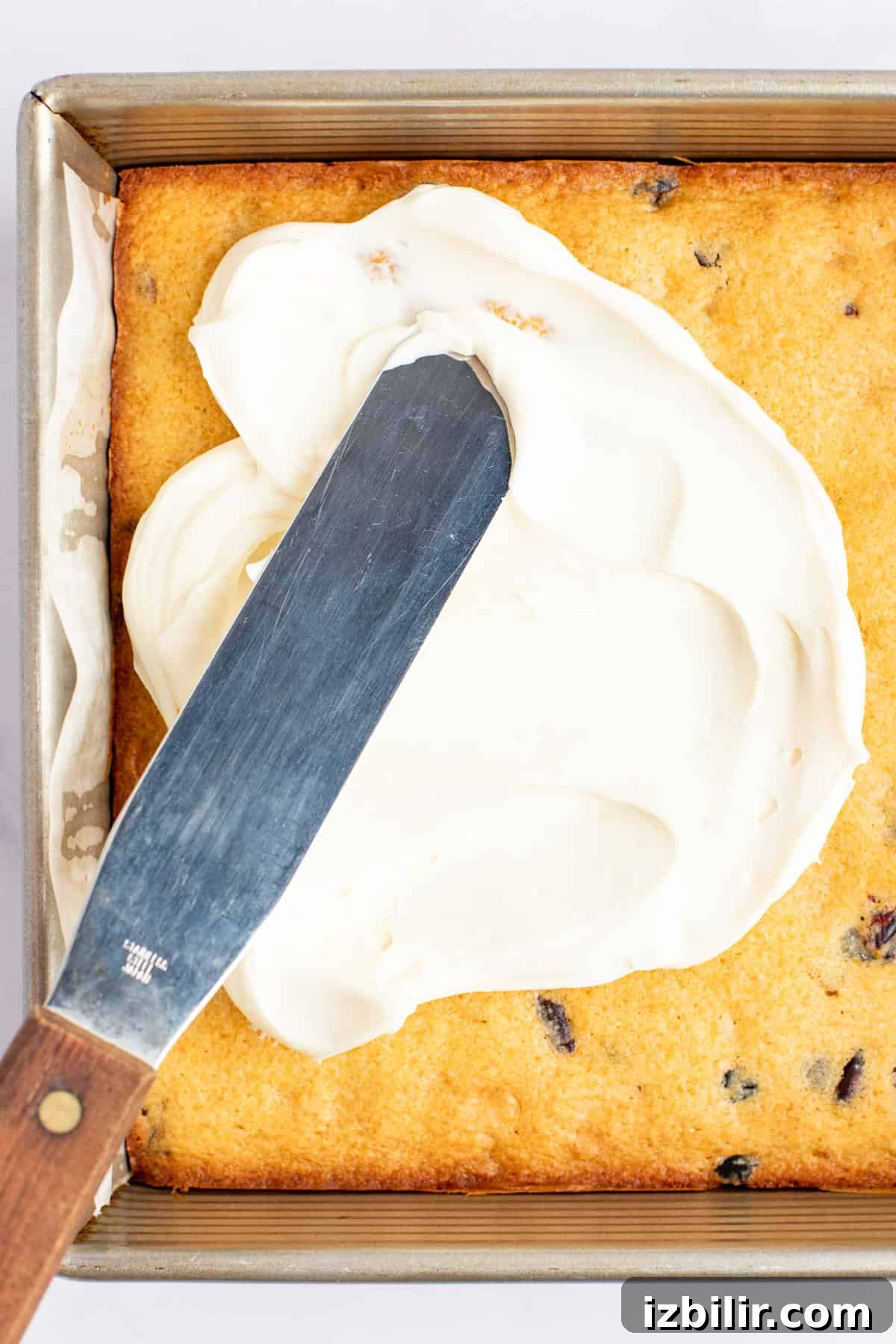 A metal spatula is spreading white frosting on a rectangular, baked cake in a square pan. The cake has visible chocolate chips.