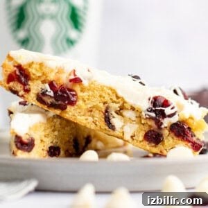 A close-up of two white chocolate cranberry bars stacked on a plate with a Starbucks cup in the background. The bars have a creamy white frosting and visible dried cranberries.