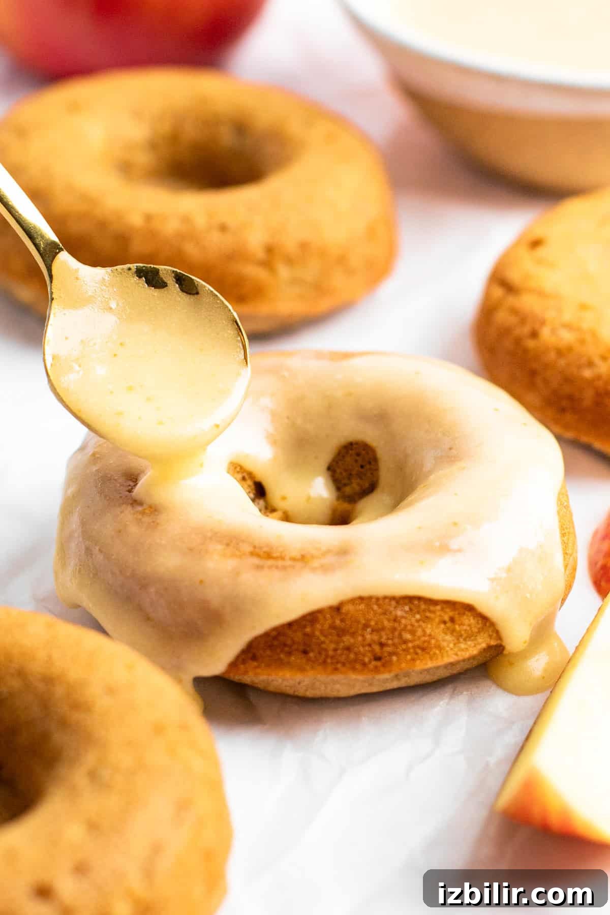 Apple Cider Donuts Recipe with Cinnamon Sugar 6 A spoon drizzles glaze over a baked donut with more donuts and a sliced apple in the background.