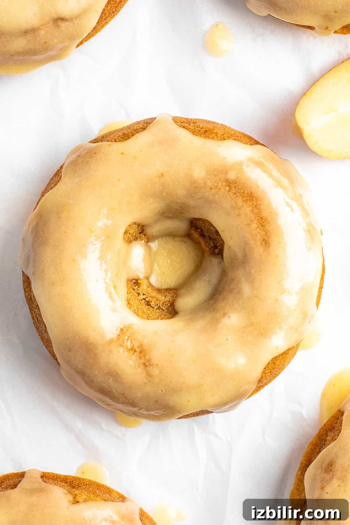 Apple Cider Donuts Recipe with Cinnamon Sugar 7 Close-up of a glazed donut with a smooth, shiny coating on a white surface. The donut has a hole in the center with some exposed texture. Another donut is partially visible in the background.