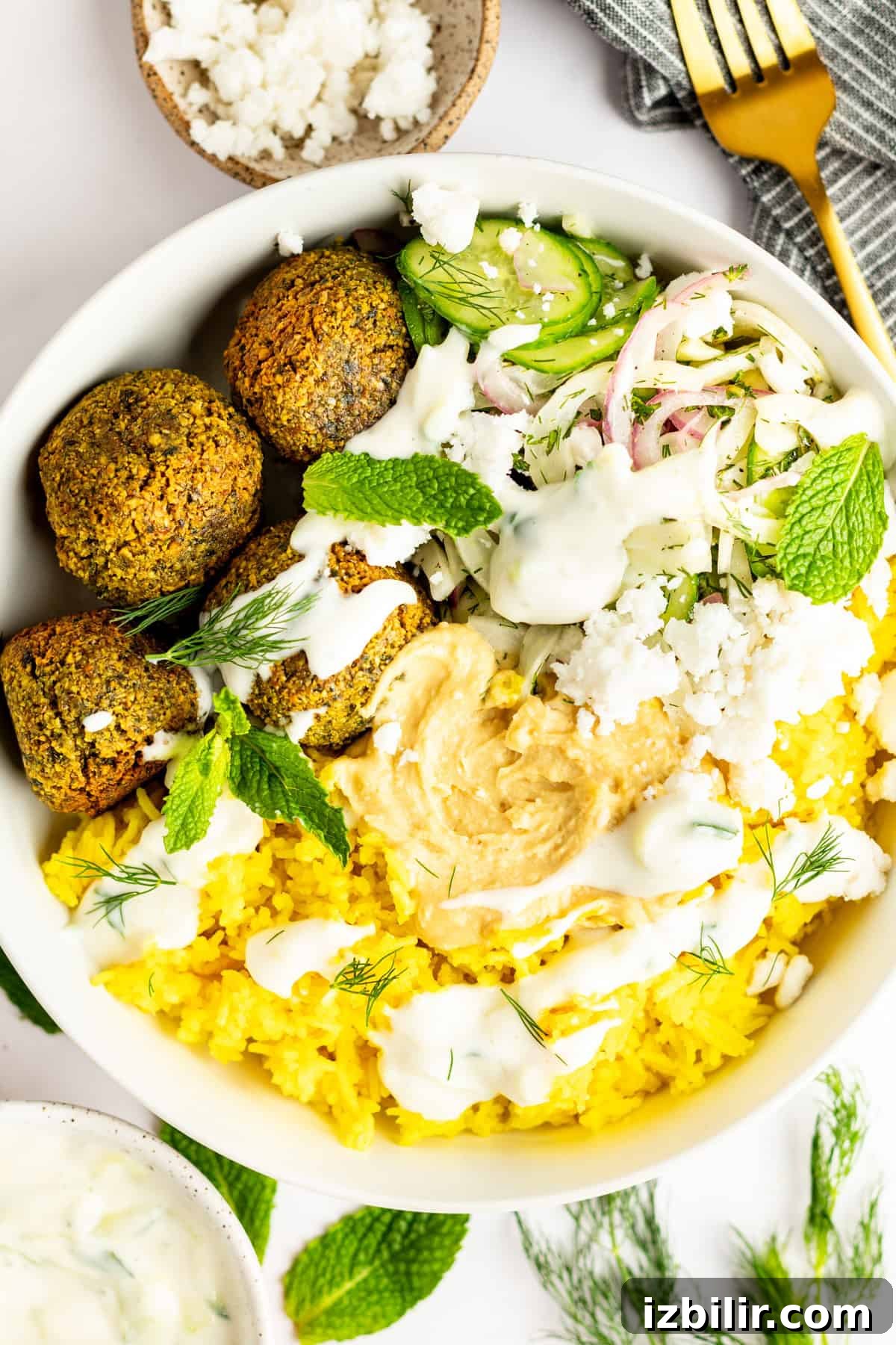 A bowl of falafel, yellow rice, cucumber salad, hummus, feta cheese, and herbs, with a drizzle of white sauce. A gold fork and a gray napkin are placed beside the bowl.