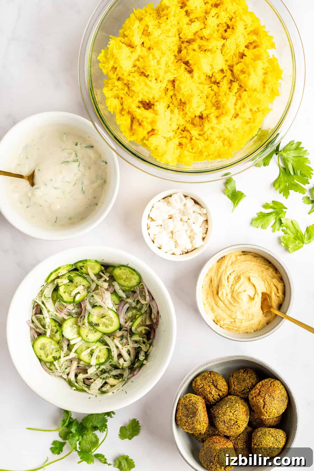 Assorted dishes including a bowl of yellow rice, yogurt sauce, feta cheese crumbles, hummus, cucumber salad, and falafel, arranged on a white surface with sprigs of cilantro.