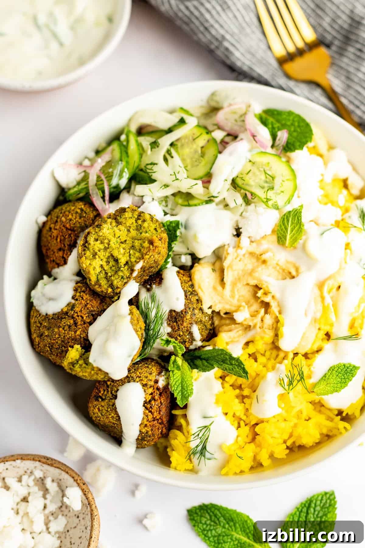 A bowl of yellow rice, falafel, sliced cucumbers, red onions, hummus, and white sauce garnished with mint leaves and herbs. A fork and a small bowl of dip are in the background.