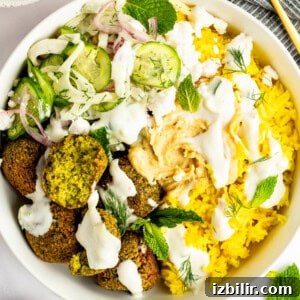 A bowl containing yellow rice, falafel, sliced cucumber, red onions, dill, feta cheese, and mint leaves, topped with hummus and drizzled with a white sauce.
