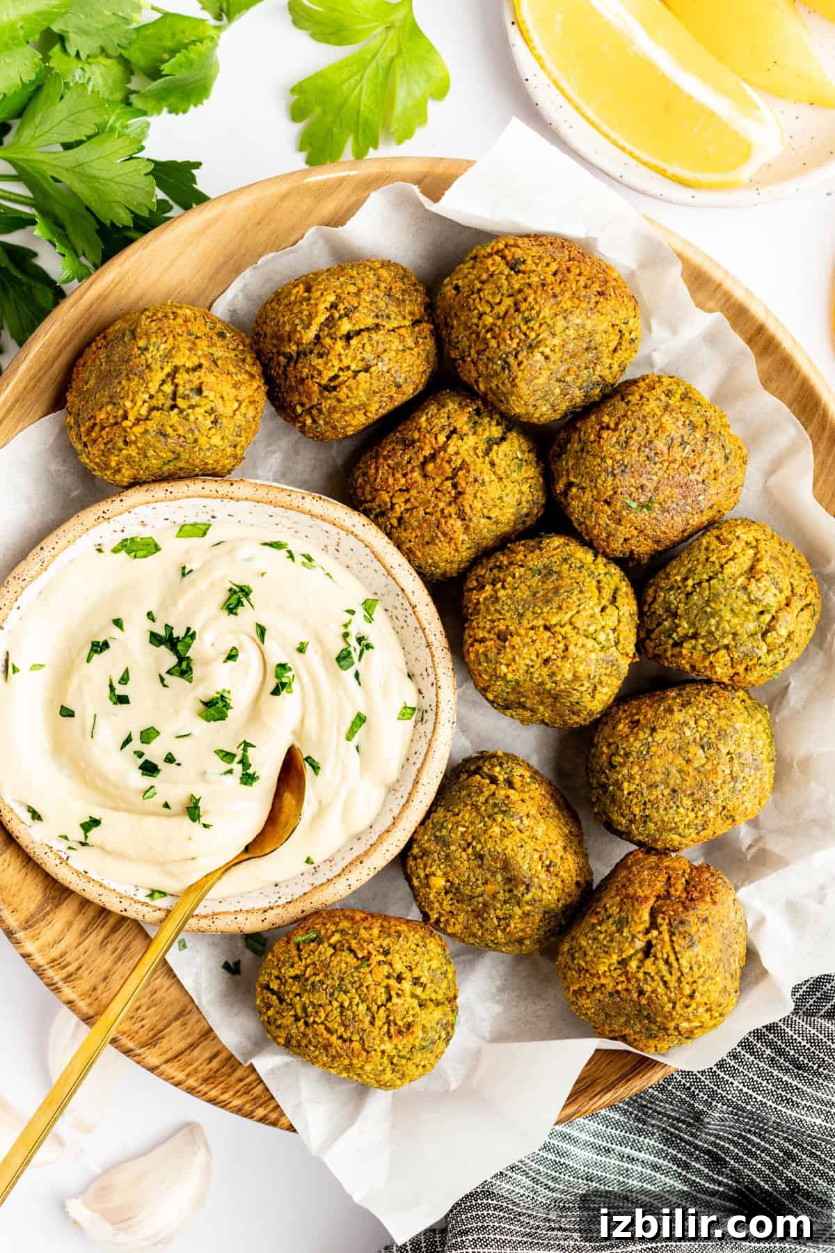 Crispy Homemade Falafel Recipe with Tahini Sauce 2 A wooden plate holds ten falafel balls surrounding a bowl of creamy dip garnished with herbs. Lemon wedges and parsley leaves are in the background.