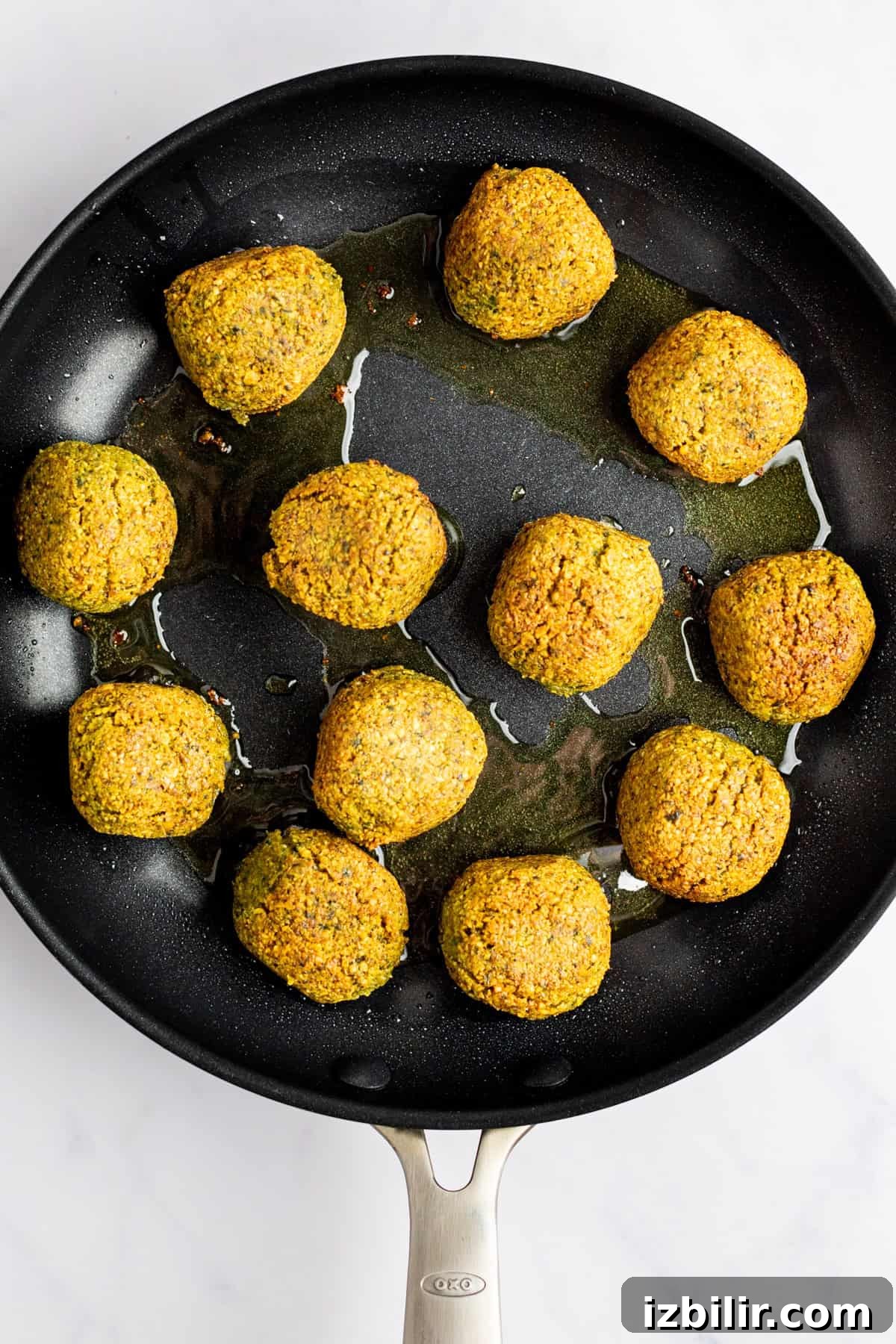 Crispy Homemade Falafel Recipe with Tahini Sauce 6 A skillet with twelve golden-brown falafel balls frying in oil on a light-colored surface.