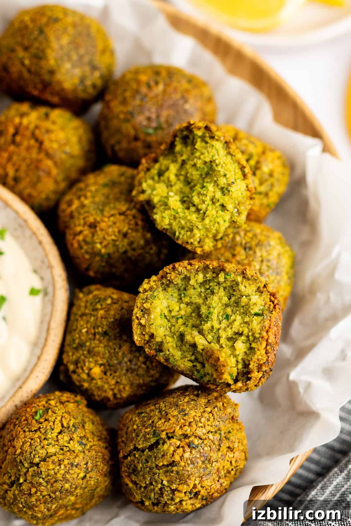 Crispy Homemade Falafel Recipe with Tahini Sauce 7 A close-up of a basket of falafel balls, with one broken open to show the green interior. A small bowl of dipping sauce is partially visible next to the falafel.