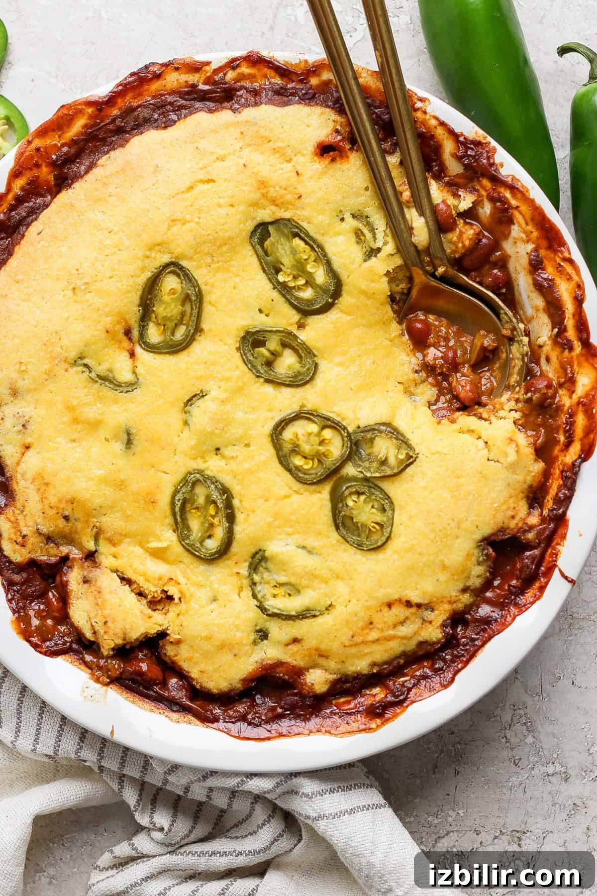 A dish of cornbread chili pie topped with sliced jalapeños, served in a white pie dish, with two gold-colored spoons placed on top, set on a light-colored surface.