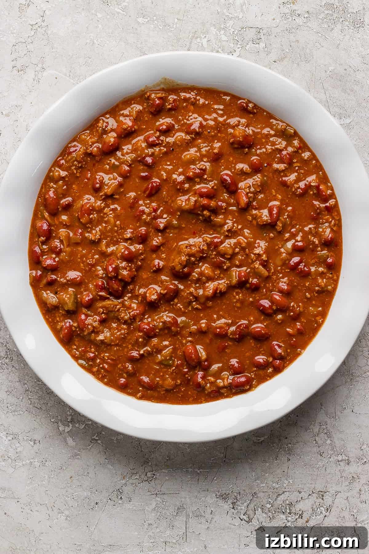 A white bowl filled with chili, containing ground meat, beans, and a thick tomato-based sauce, placed on a textured gray surface.