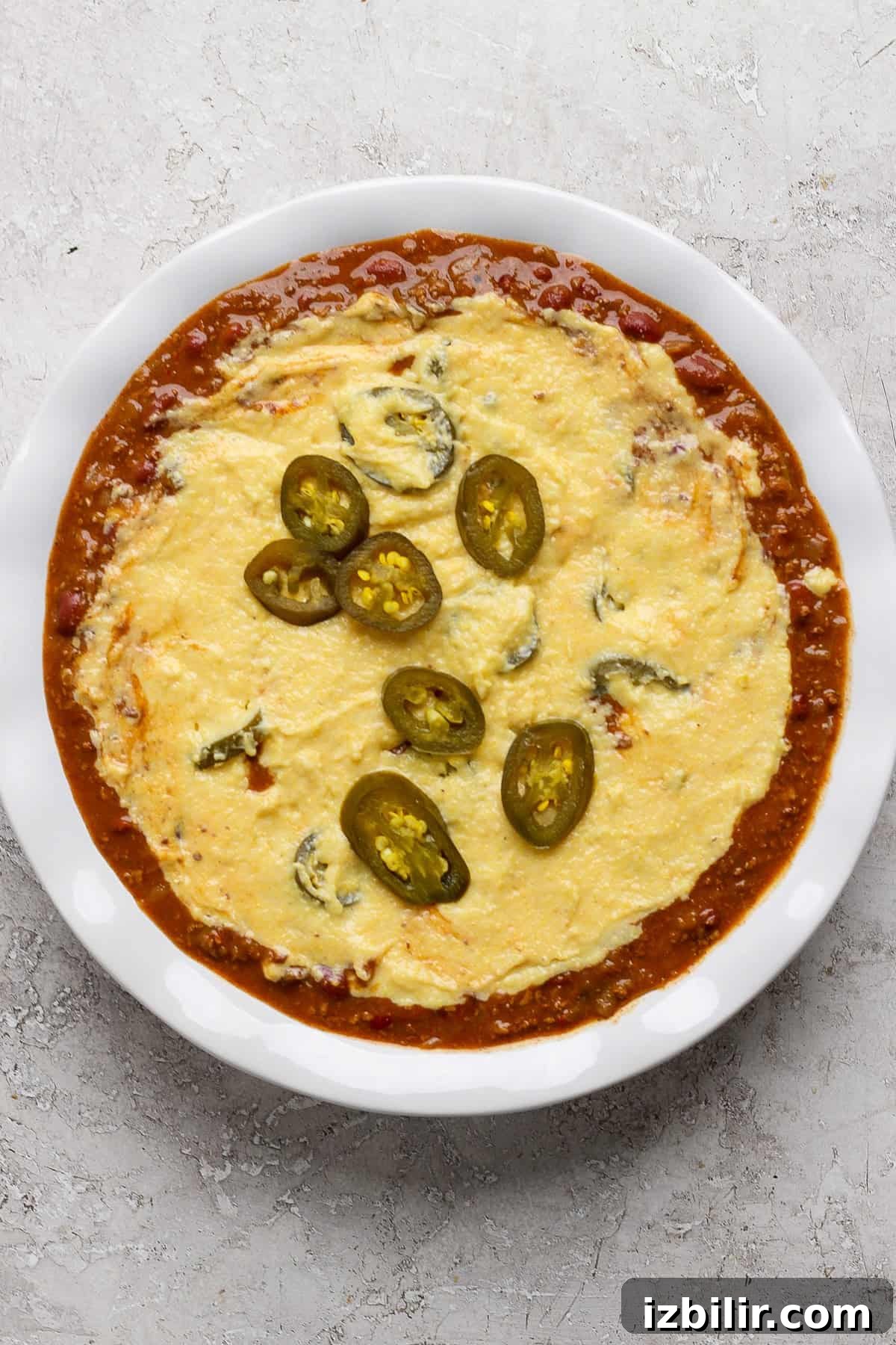 A white dish filled with melted cheese topped with sliced jalapeños, surrounded by a red tomato-based sauce.