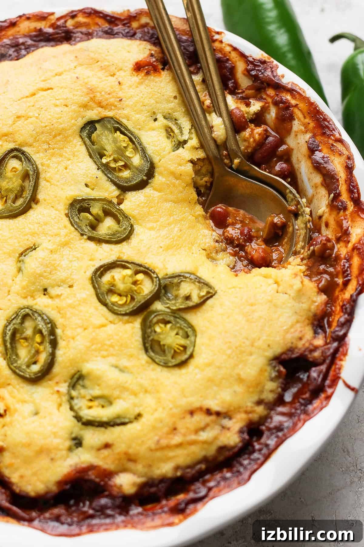 A baked dish in a white pie dish features a golden cornbread topping with sliced jalapeños, partially revealing a chili filling underneath. Two metal spoons are placed in the dish.