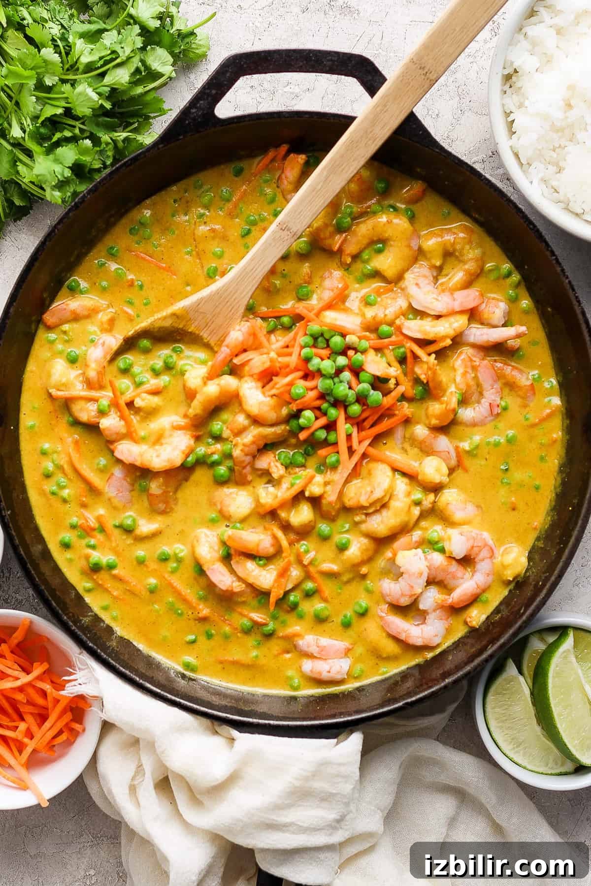 One-Pot Trader Joe's Yellow Curry Recipe 2 A skillet filled with shrimp and vegetable curry, garnished with green peas and served with sides of rice, lime, and fresh herbs.