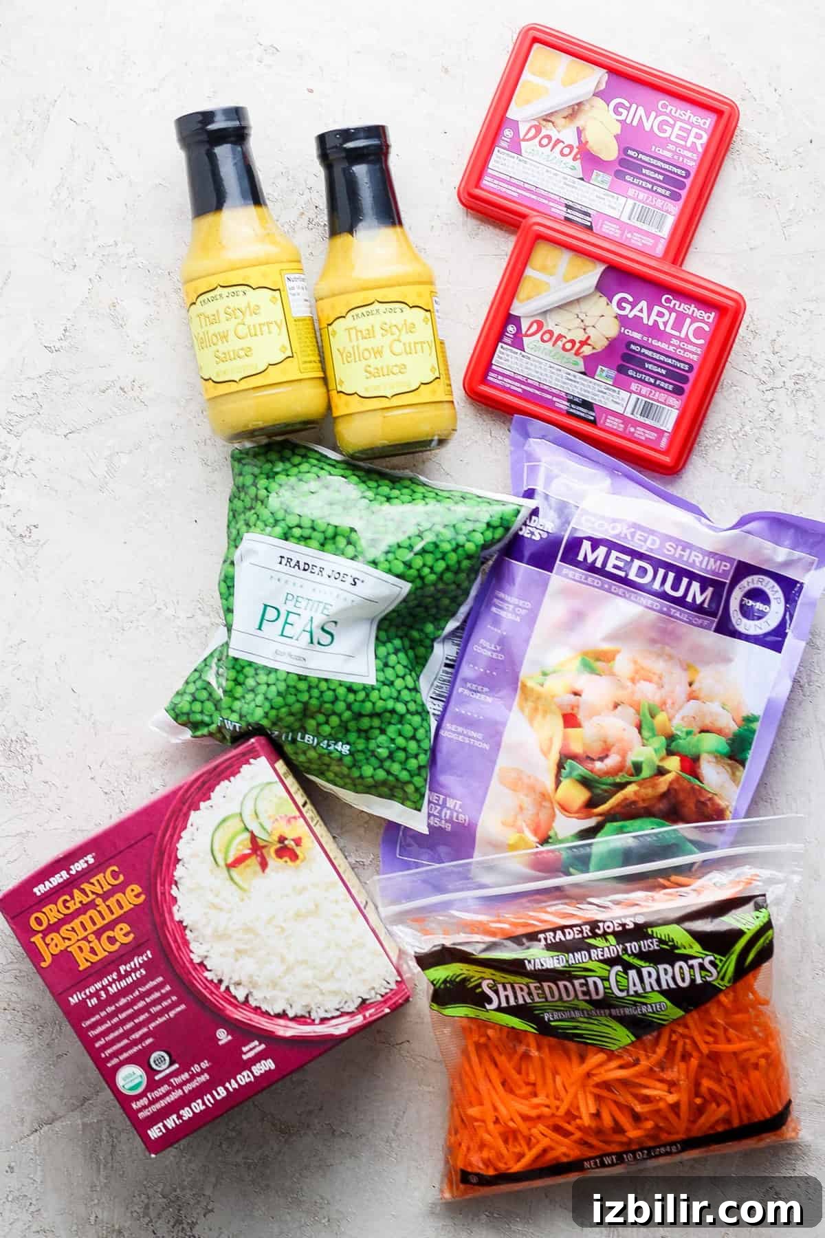 One-Pot Trader Joe's Yellow Curry Recipe 3 A collection of various food items, including bottles of thai yellow curry sauce, a box of ginger, packaged garlic, organic jasmine rice, frozen peas, and shredded carrots, arranged on a light textured surface.