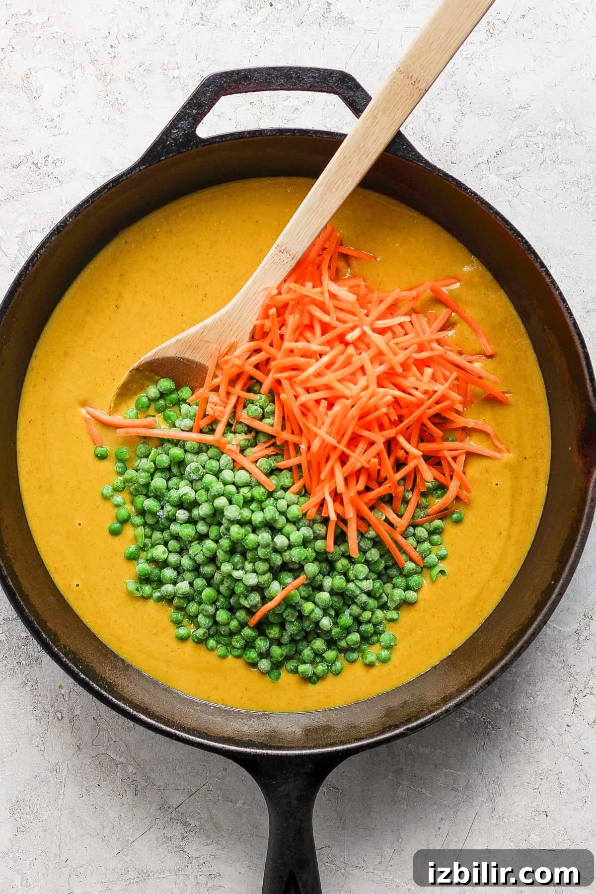 One-Pot Trader Joe's Yellow Curry Recipe 4 A cast iron skillet containing creamy soup garnished with a spoonful of green peas and a scattering of grated carrots on a light-colored surface.