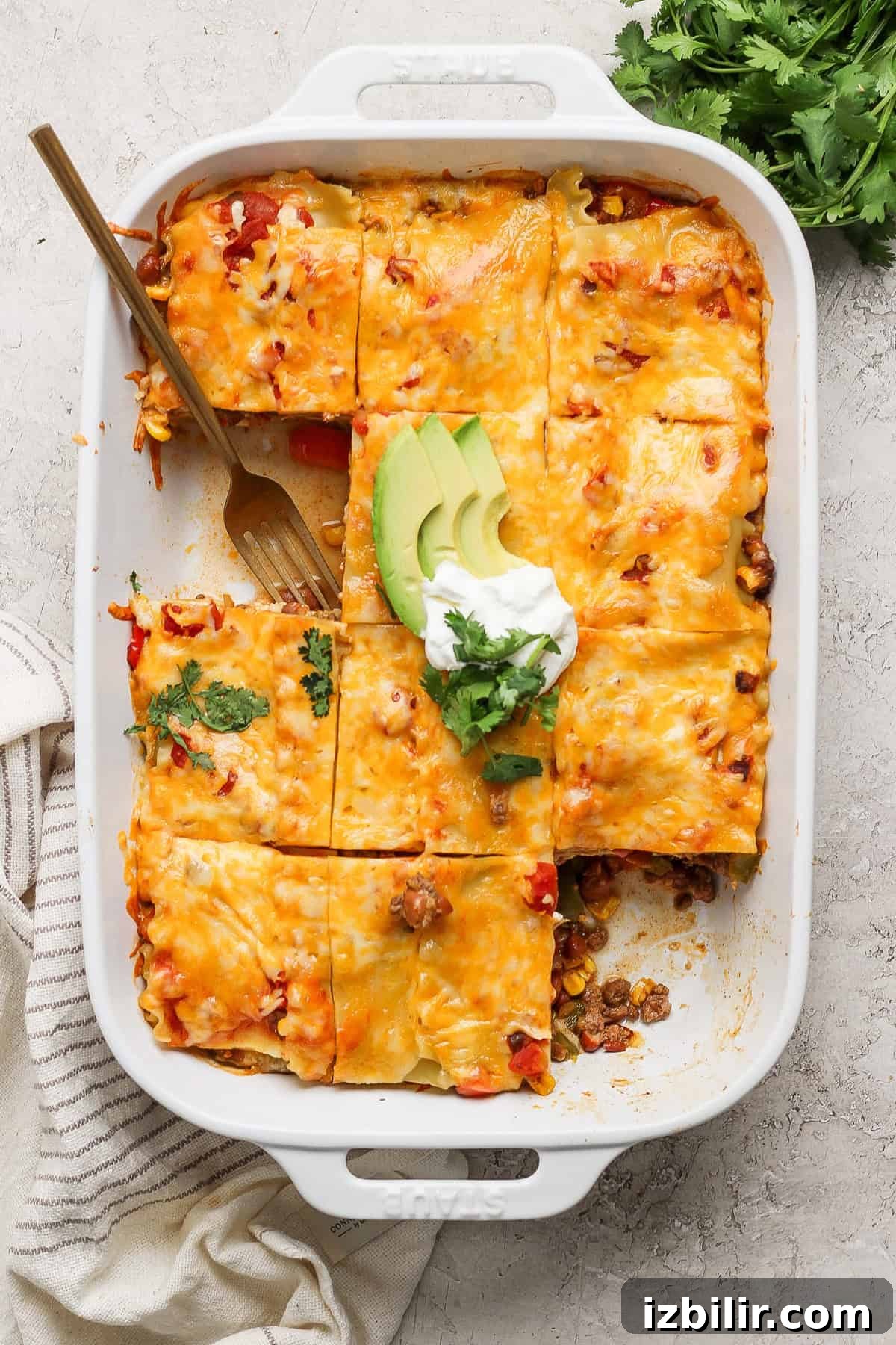 A baked casserole in a white rectangular dish divided into squares topped with melted cheese, garnished with avocado slices, sour cream, and cilantro.