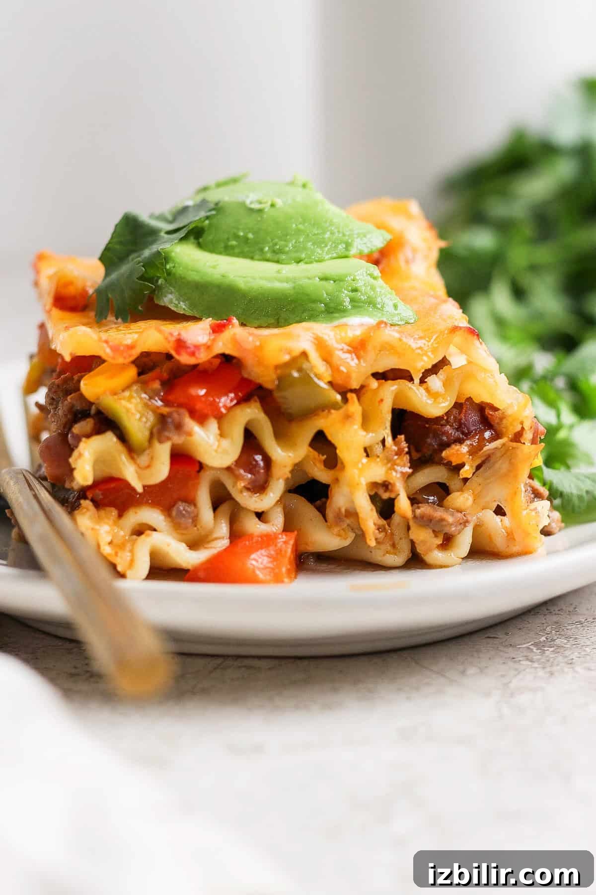 A close-up of a slice of taco lasagna topped with avocado slices and herbs on a white plate.