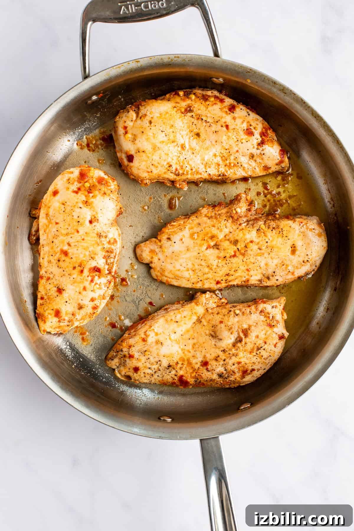 Four seasoned chicken breasts are cooking in a stainless steel skillet.