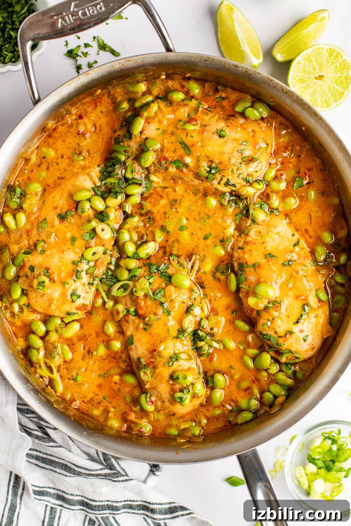 A stainless steel pan filled with cooked fish fillets in a vibrant orange sauce, garnished with chopped herbs and edamame beans. Lime wedges and a striped kitchen towel are placed beside the pan.