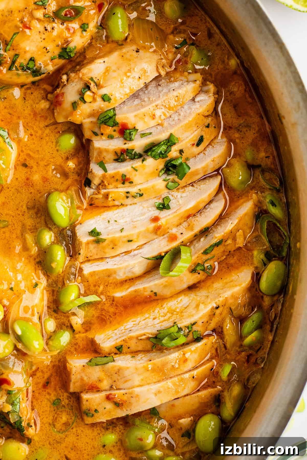 Close-up of sliced cooked chicken in a pan with a creamy, orange-colored sauce, garnished with chopped herbs and green peas.
