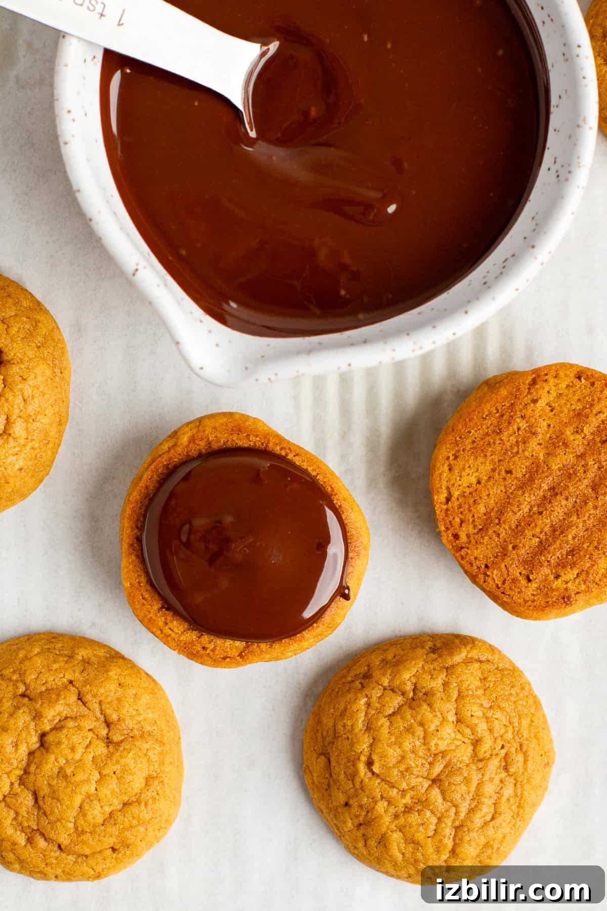 Cookies being dipped in melted chocolate from a white bowl, with additional cookies placed nearby on a white surface.