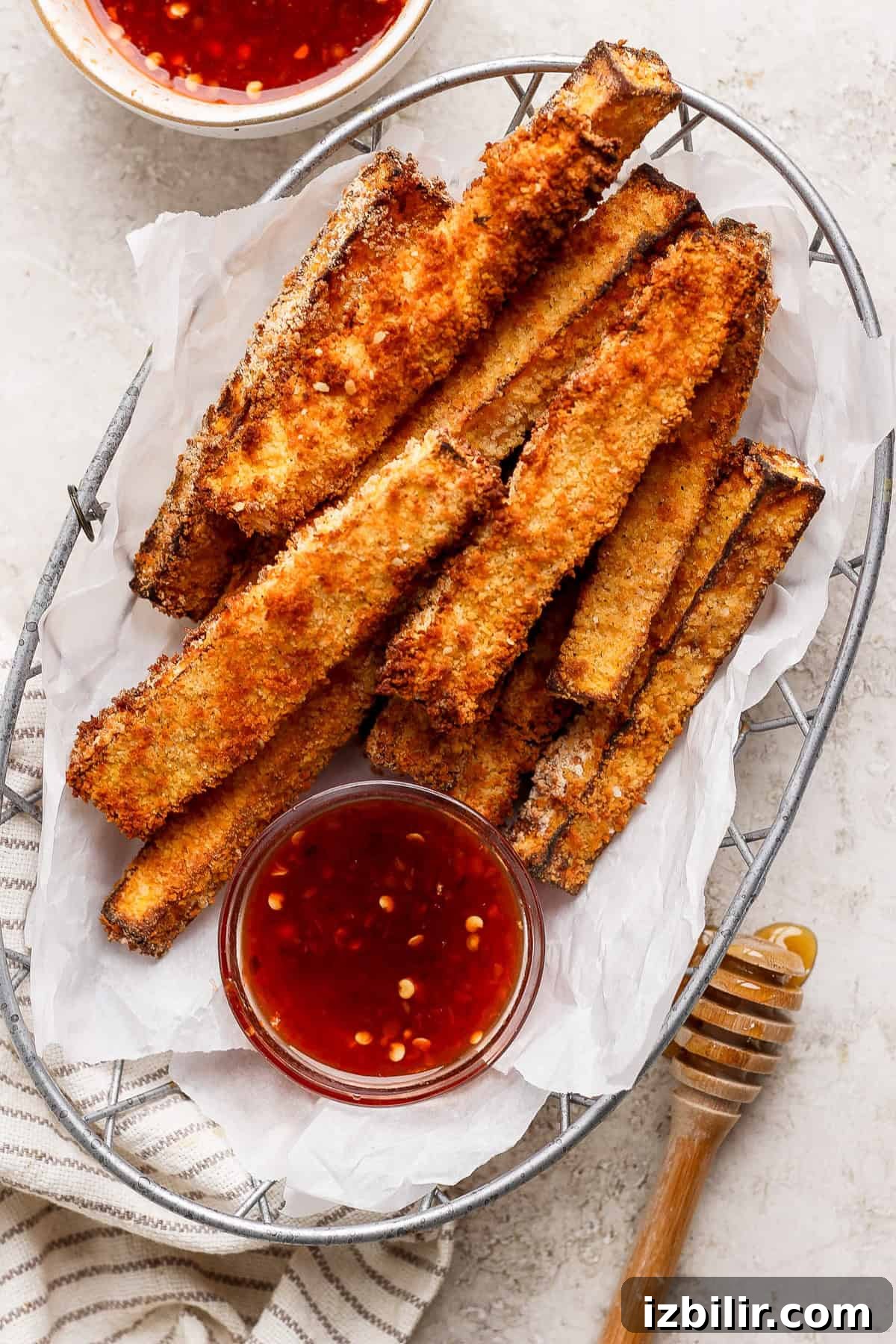 Air Fryer Butternut Squash Fries with Hot Honey Recipe 2 A basket of crispy, breaded zucchini fries served with two small bowls of red dipping sauce and a wooden honey dipper on a striped cloth.
