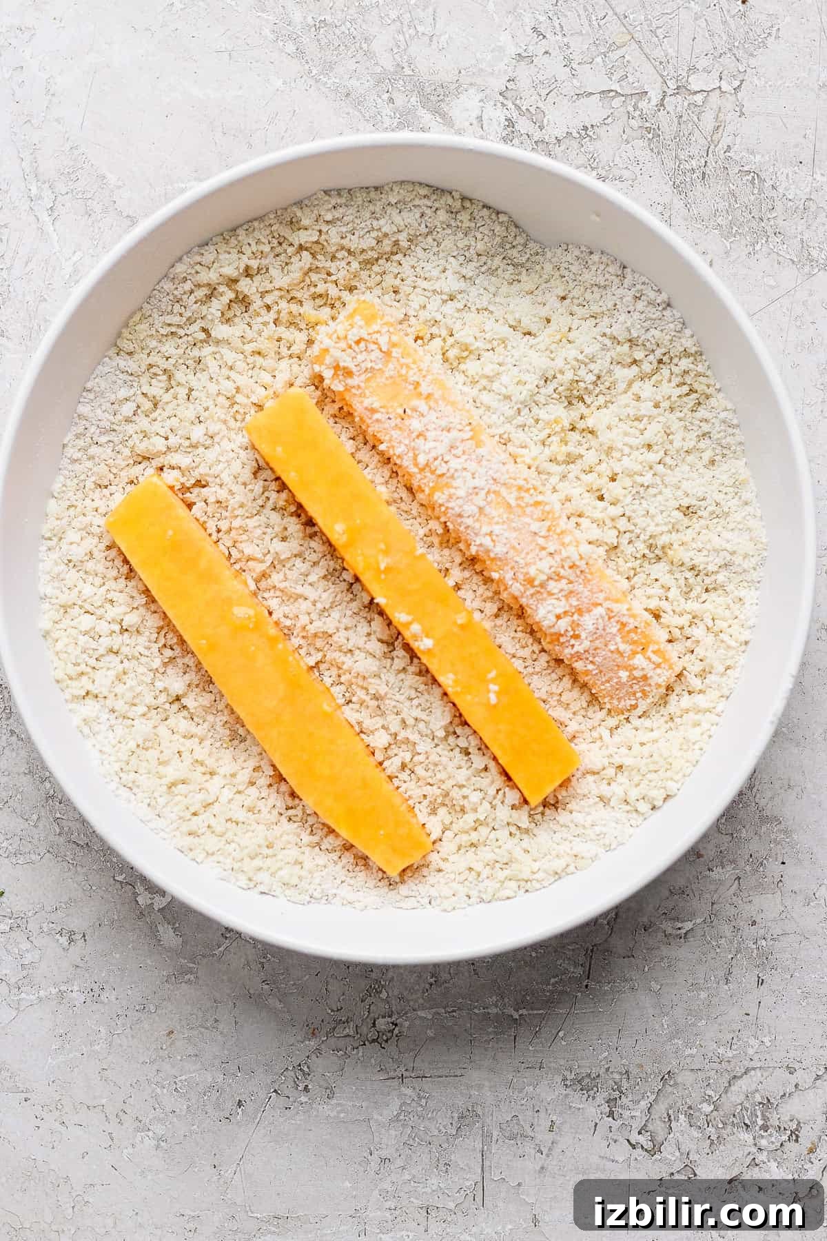 Air Fryer Butternut Squash Fries with Hot Honey Recipe 3 Three breaded cheese sticks are positioned in a bowl filled with breadcrumbs on a textured white surface.