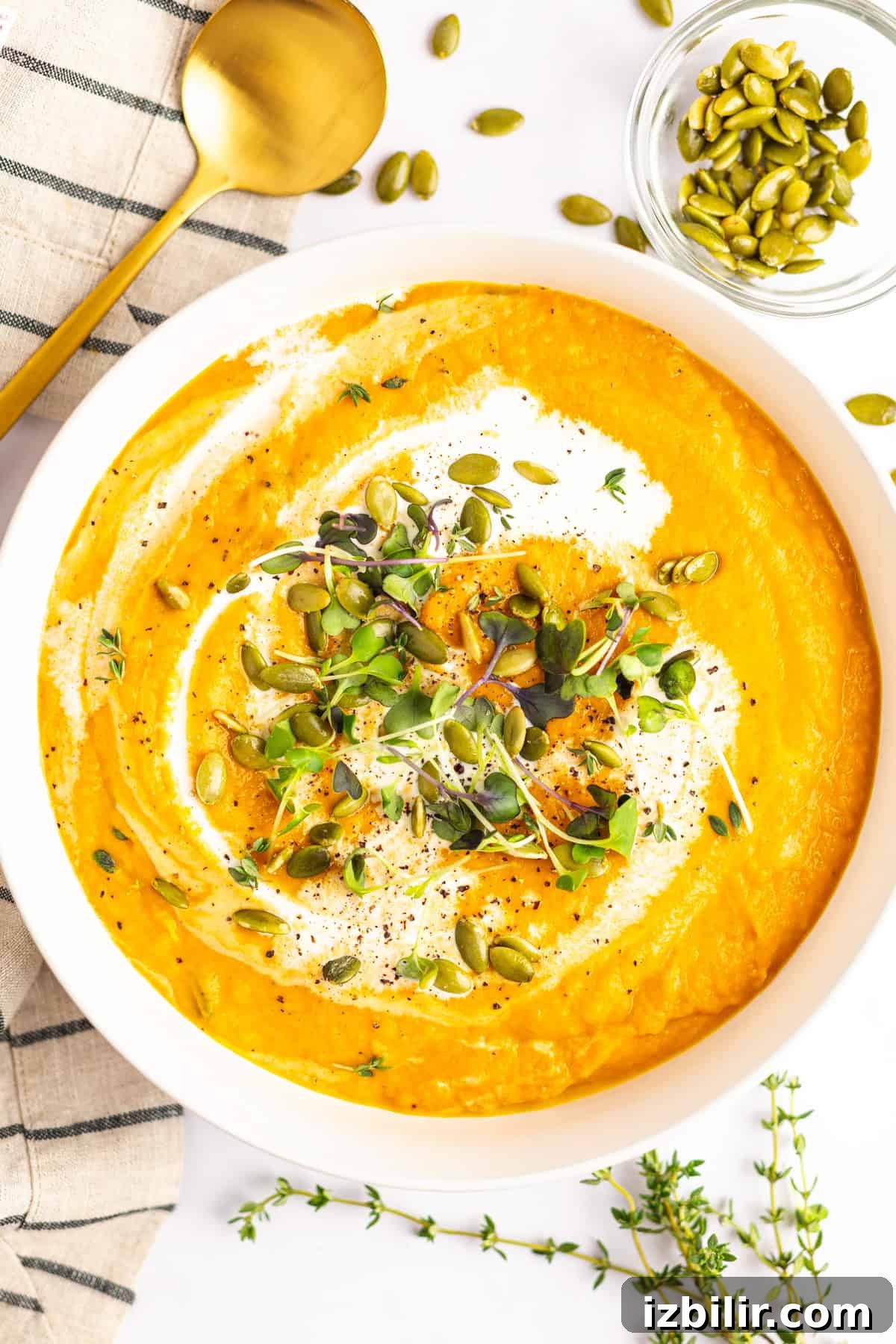 A bowl of creamy orange soup garnished with microgreens, pumpkin seeds, and a swirl of cream. A gold spoon, sprigs of fresh herbs, and a small bowl of pumpkin seeds are placed nearby.