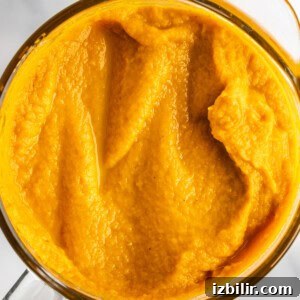 A top view of a glass container filled with smooth, bright orange pumpkin puree on a white marble surface.