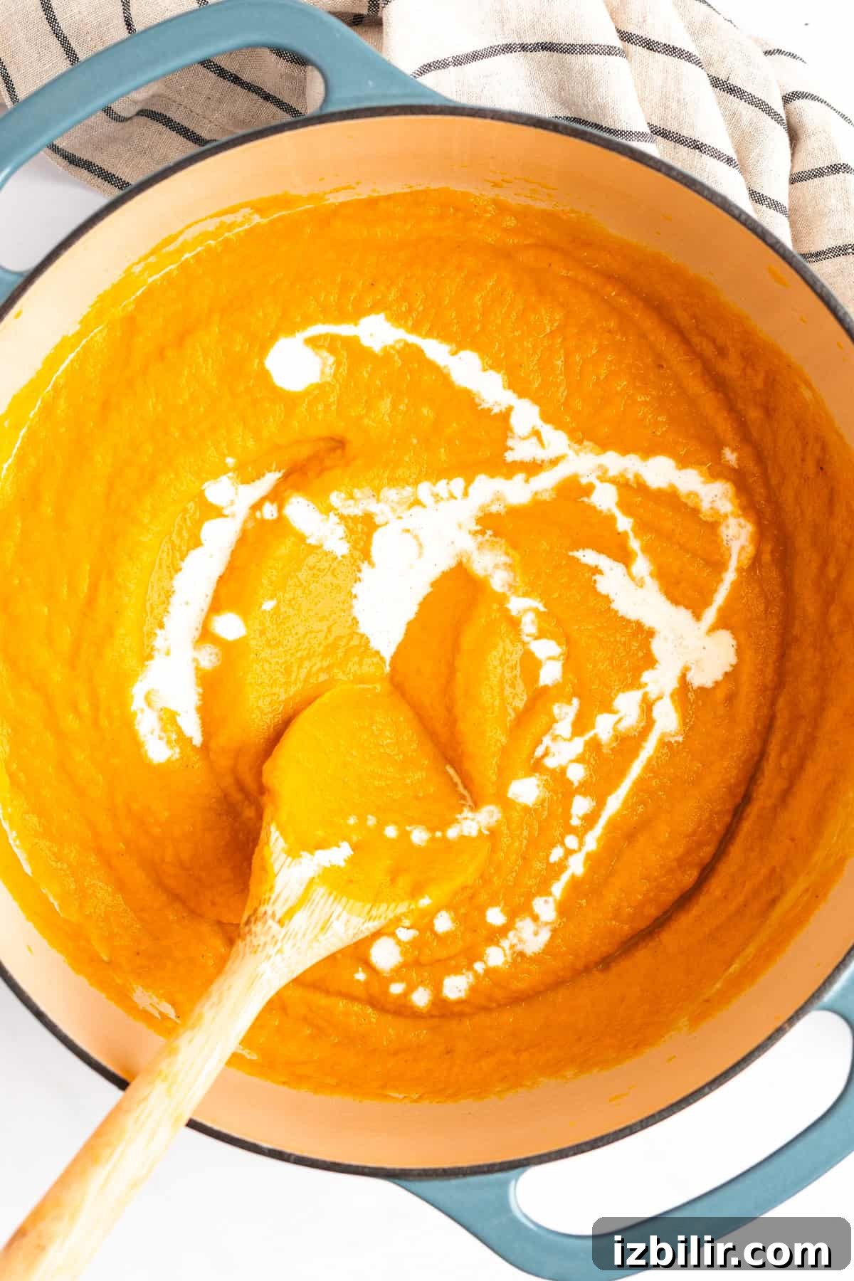 A pot of creamy orange soup is being stirred with a wooden spoon. A swirl of cream is visible on top of the soup. A striped cloth is beside the pot.