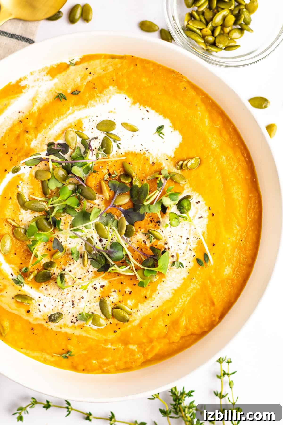 A bowl of creamy pumpkin soup garnished with a swirl of cream, microgreens, and pumpkin seeds, with additional pumpkin seeds and thyme sprigs on the side.