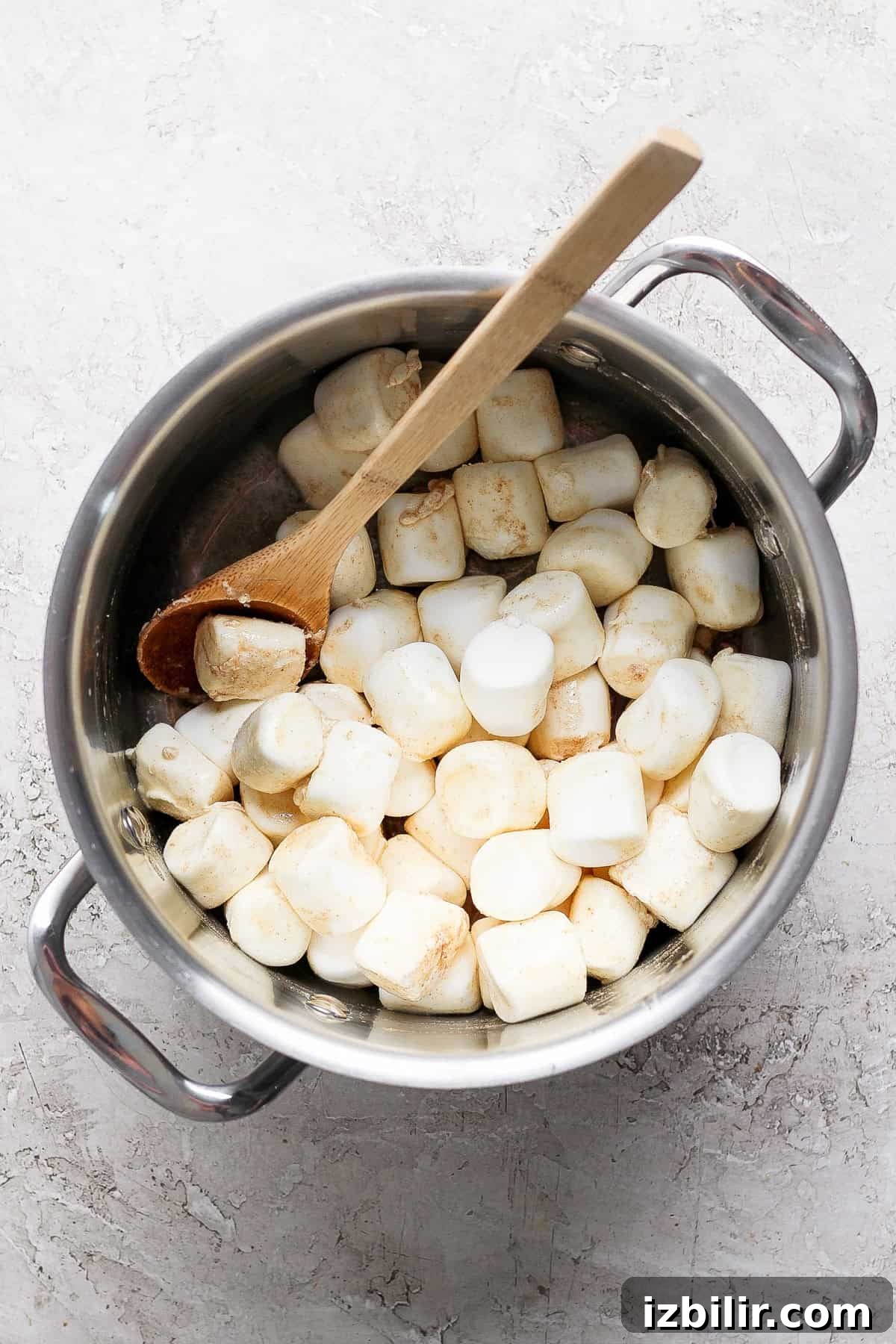 Brown Butter Chai Rice Krispie Treats Recipe 3 A pot filled with large marshmallows and a wooden spoon placed on a textured surface.