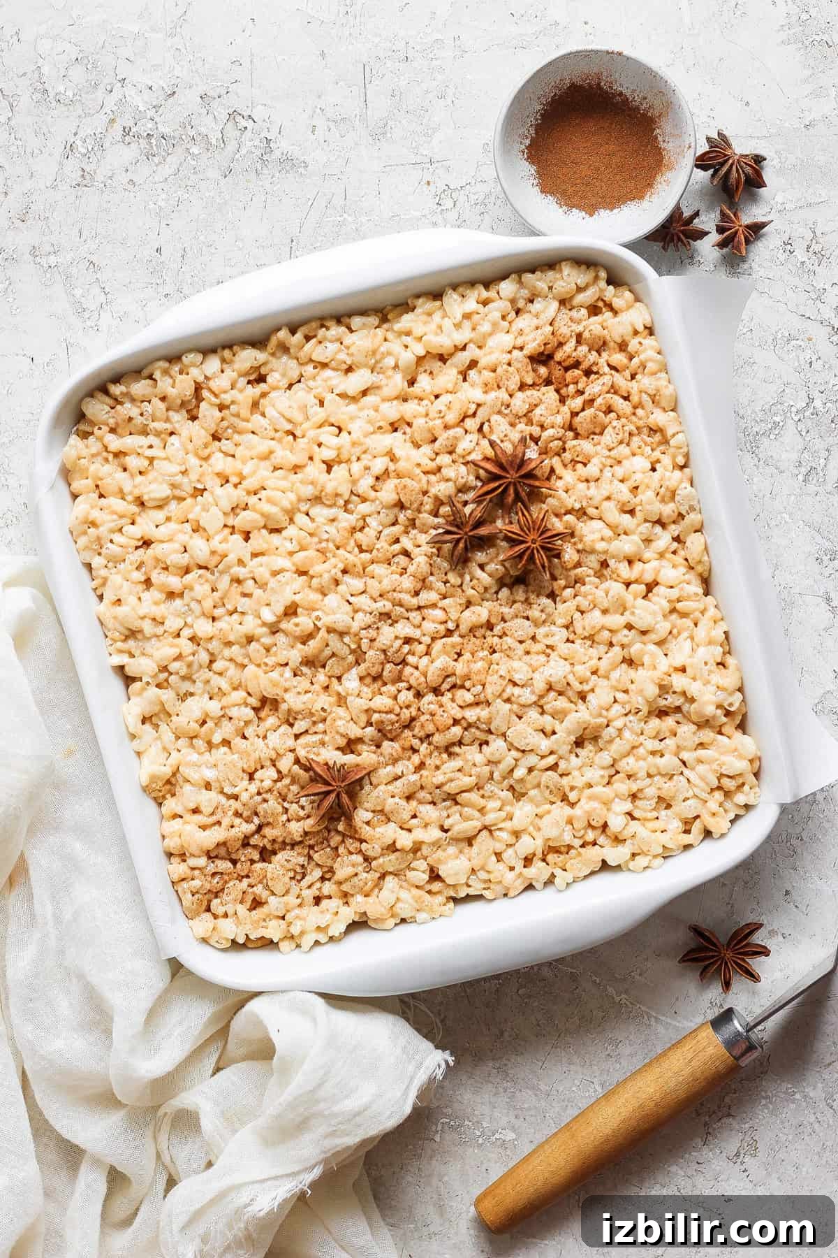 Brown Butter Chai Rice Krispie Treats Recipe 5 A square white baking dish filled with rice crispy treats, garnished with star anise and cinnamon powder in the top right corner. A small bowl with cinnamon powder is beside the dish on the counter.