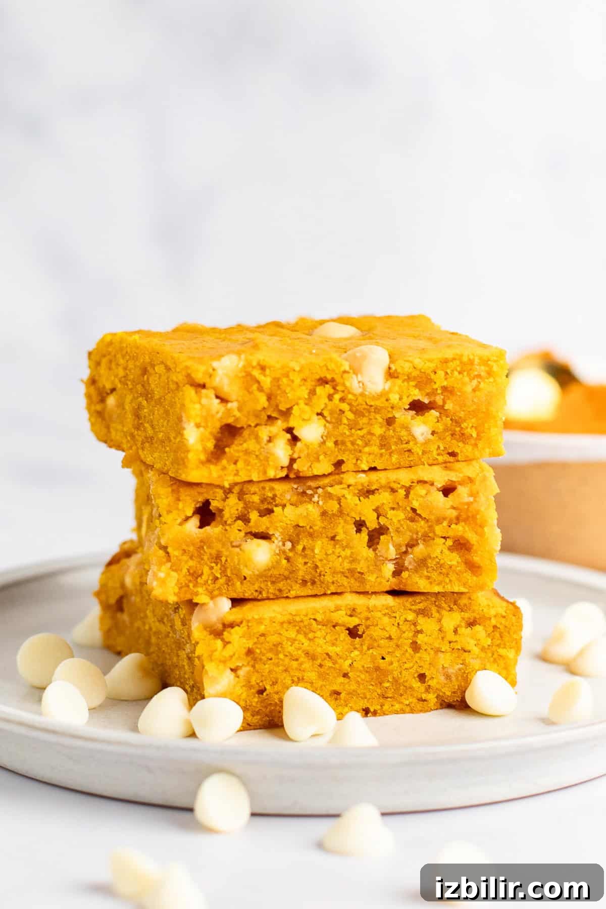 Chewy Pumpkin Blondies Recipe with Spiced Brown Sugar 2 Three slices of golden yellow pumpkin blondies stacked on a plate, surrounded by white chocolate chips.