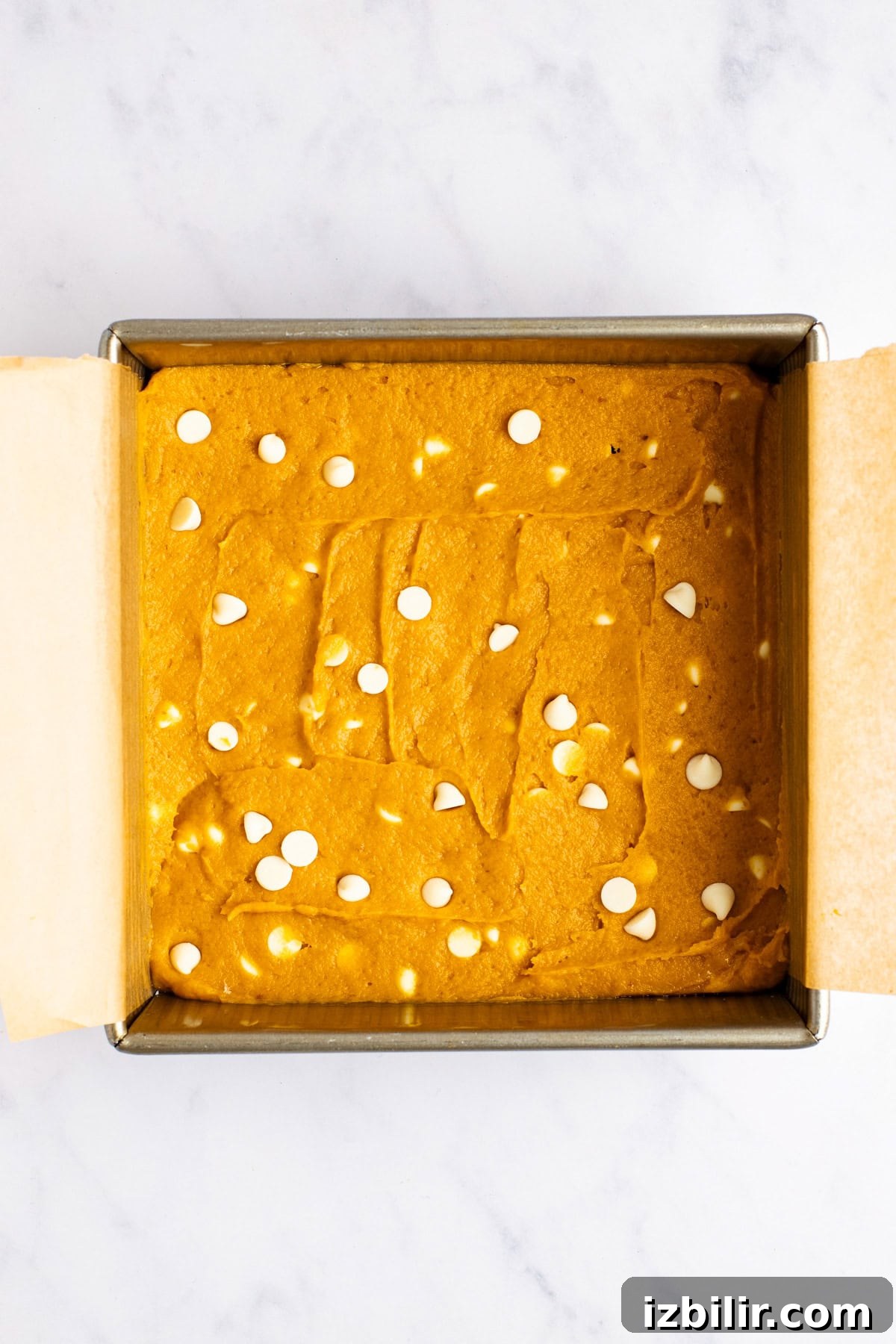 Chewy Pumpkin Blondies Recipe with Spiced Brown Sugar 3 A square baking pan filled with unbaked batter topped with scattered white chocolate chips, lined with parchment paper on a marble countertop.